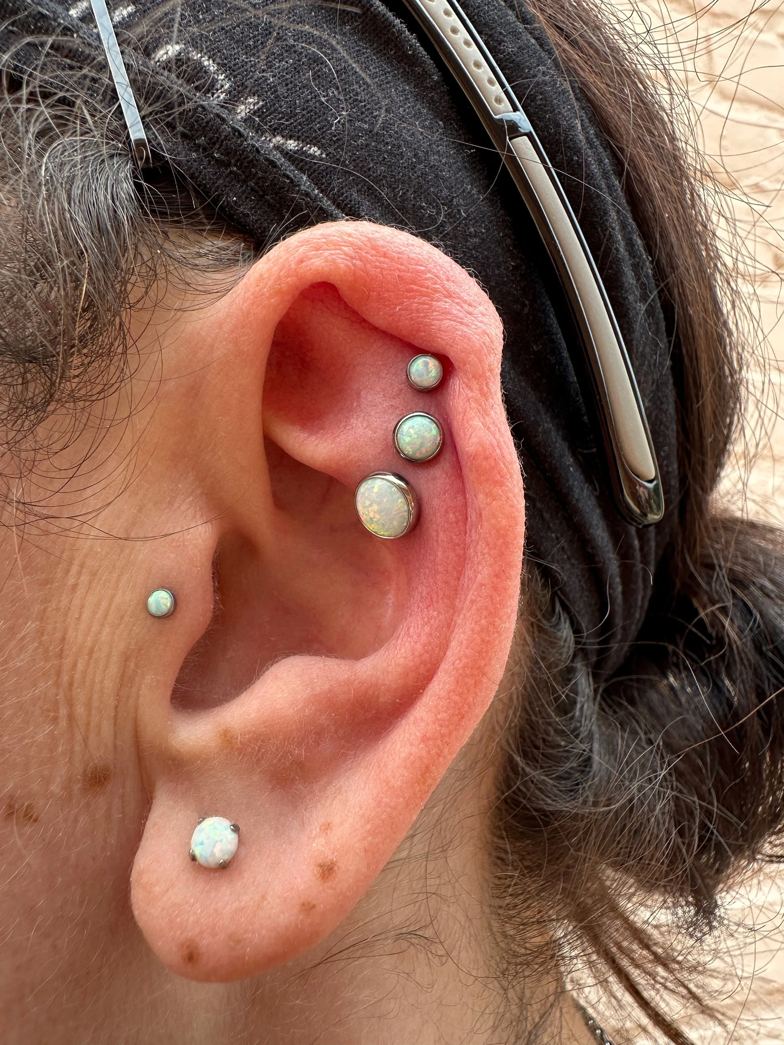 Close-up of a person's ear with five opal stud earrings in different positions. The person has dark hair and is wearing glasses.