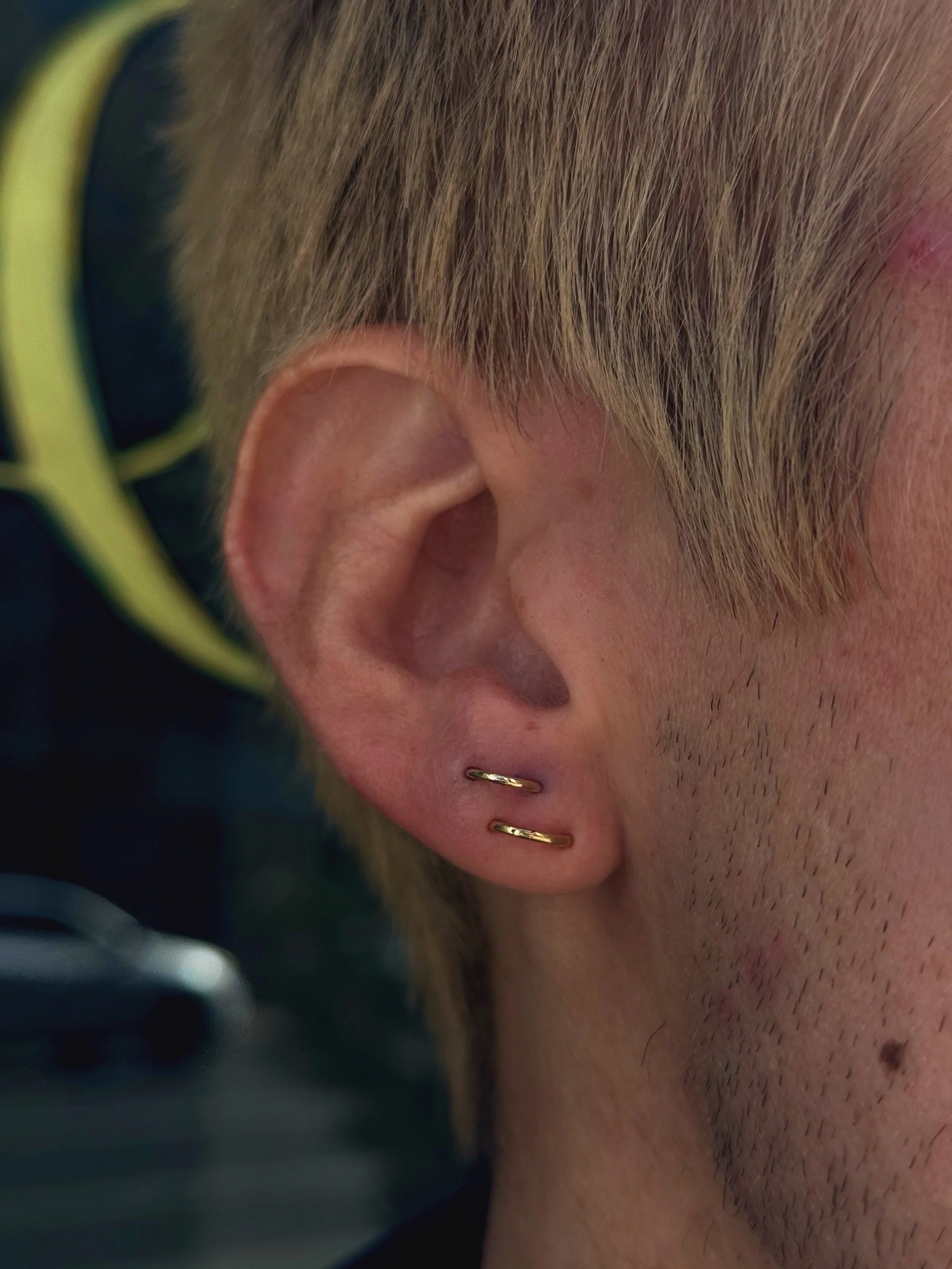 Close-up of a person's ear with two small gold hoop earrings, with blond hair and visible stubble on the face.