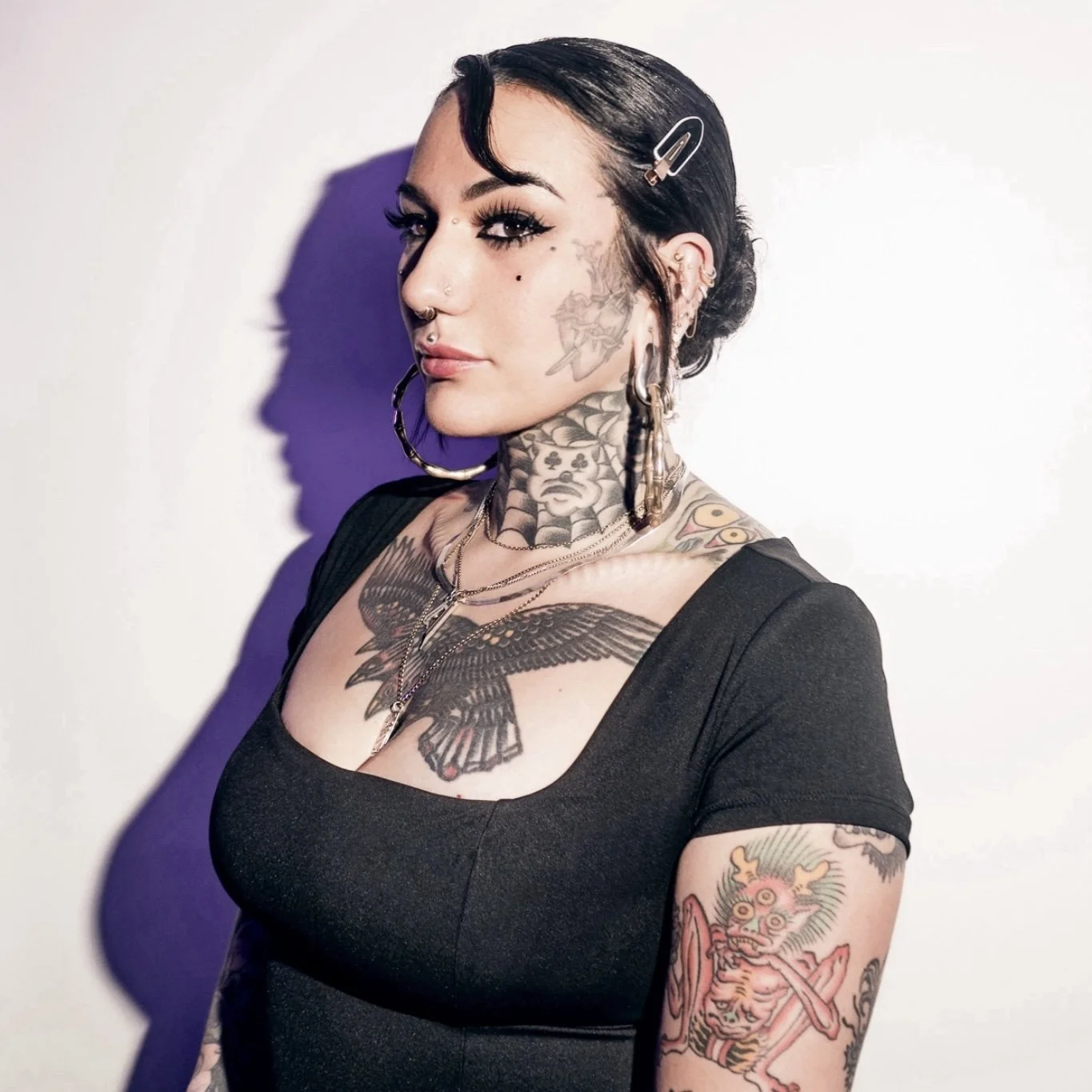 A woman with dark hair styled with a hair clip, multiple tattoos on her neck, chest, and arms, wearing large hoop earrings, and a black top, standing in front of a white wall with a shadow cast behind her.