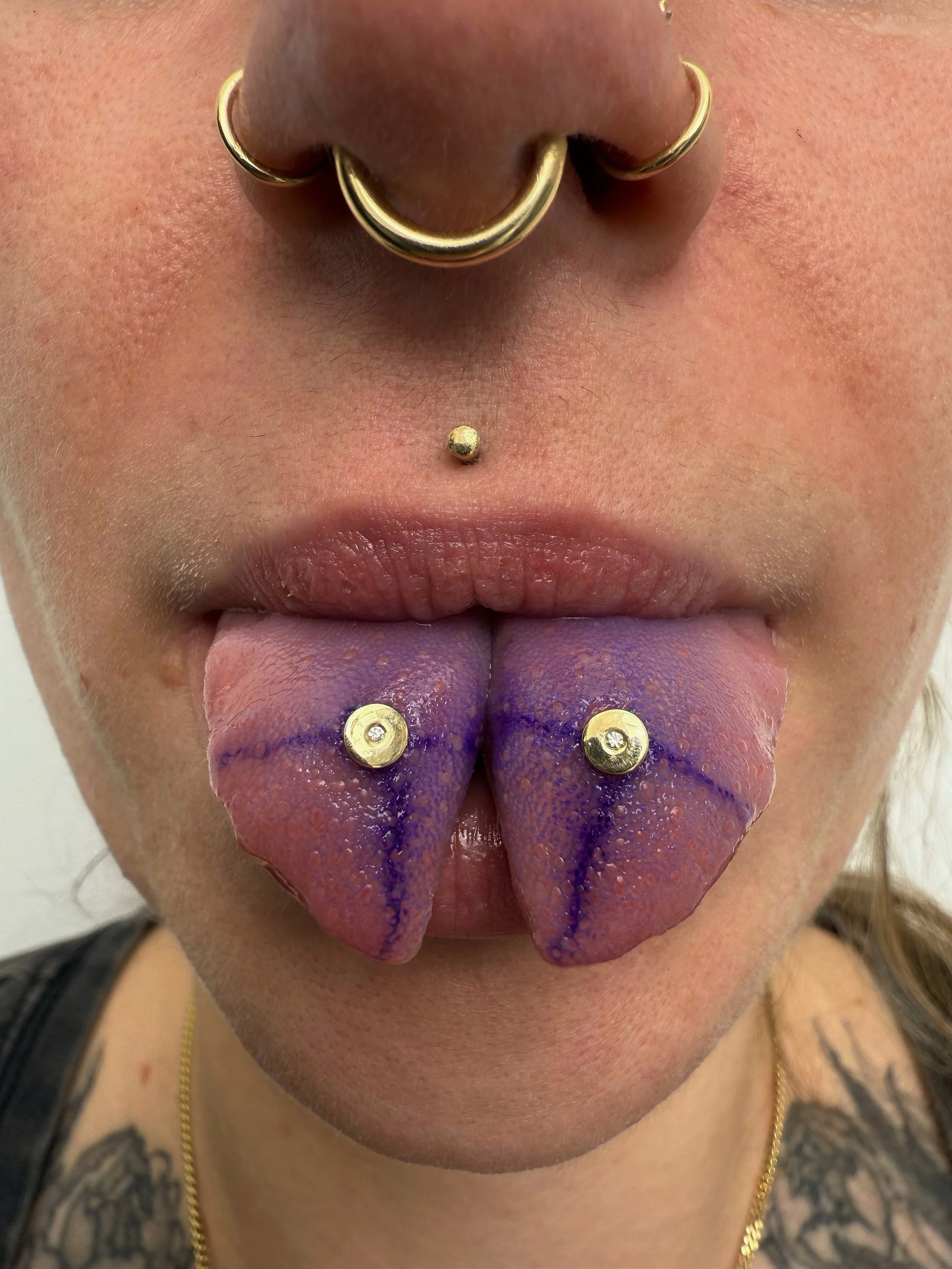 Close-up of a person's mouth with an extended tongue painted purple with dark blue lines, tongue piercings with gold balls, a septum piercing with gold hoops, and a labret piercing.