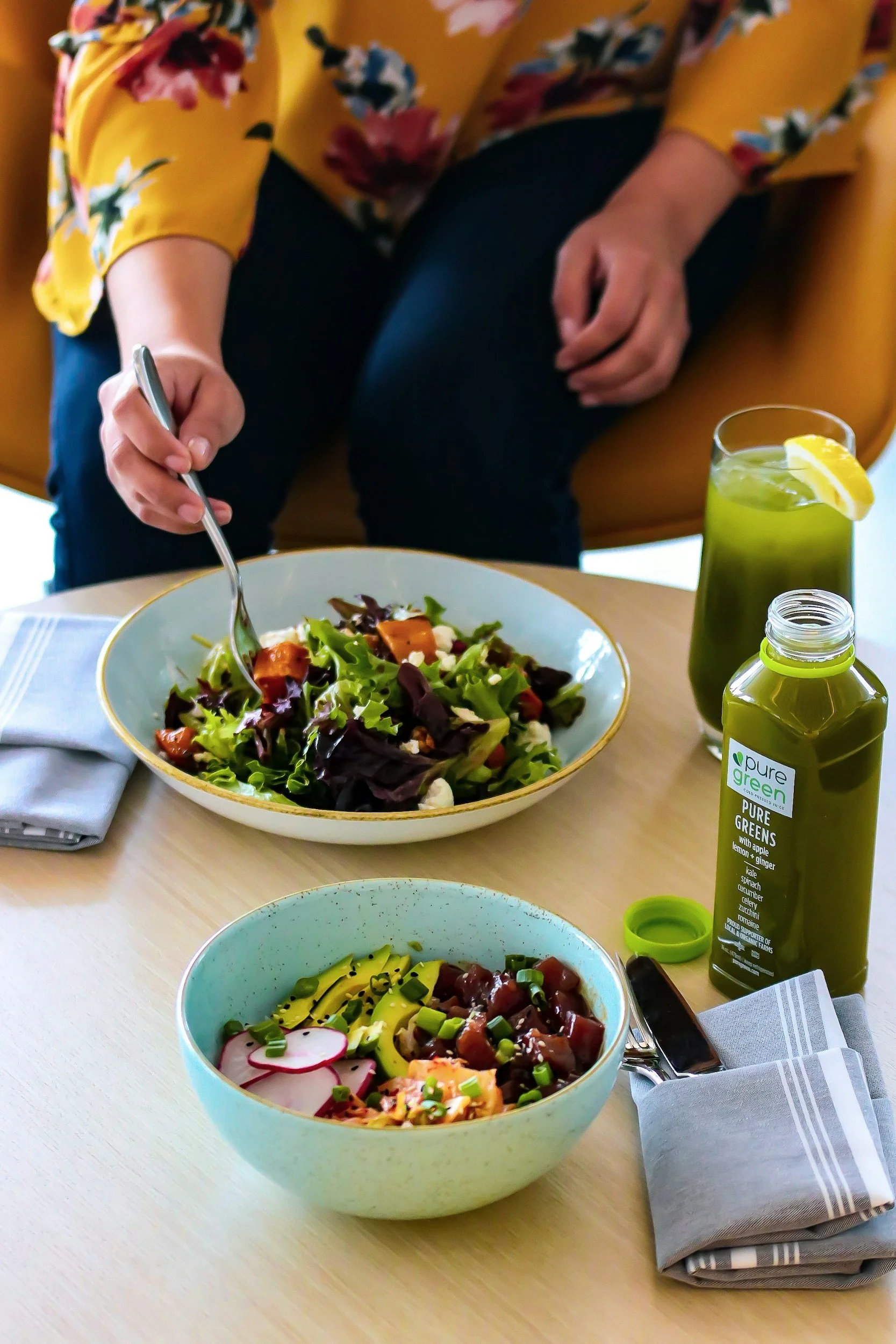 A person wearing a yellow floral blouse is preparing a salad at a table with two bowls of salad, a bottle of green juice, and a glass of green beverage with a lemon slice.
