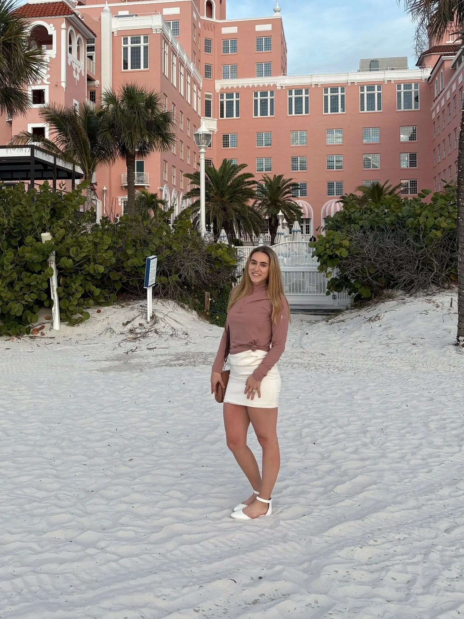 A woman with long blonde hair standing on sandy beach with a pink hotel building, palm trees, and white umbrellas in the background.