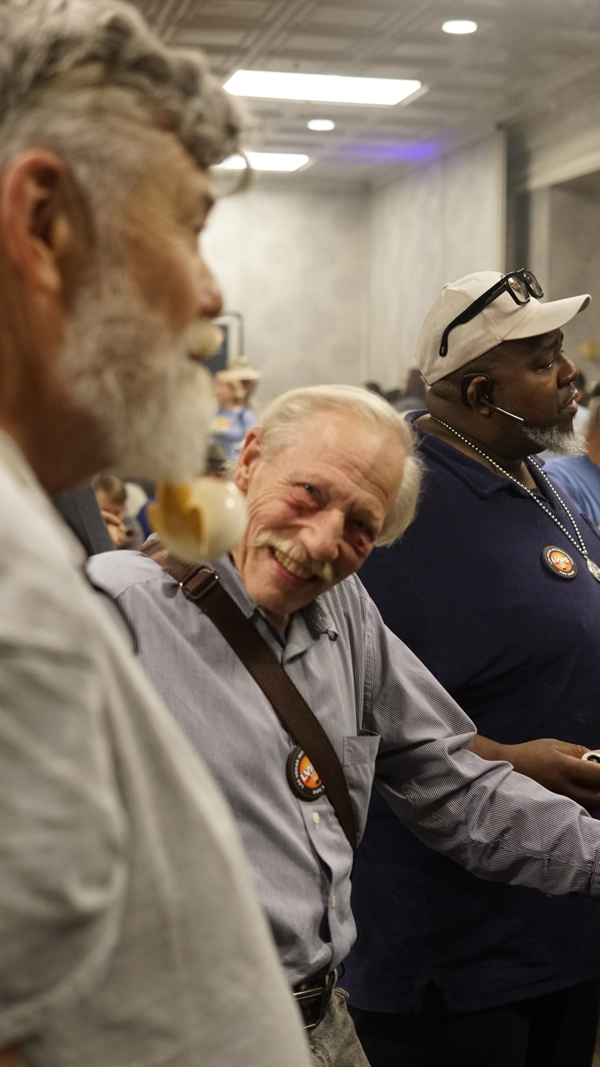 Three men sitting in a row at an indoor event, smiling and engaging. The man in the middle has white hair and a mustache, wearing a light purple shirt with a political button, and appears to be enjoying himself. The man on the left has a beard, glasses hanging from his collar, and a sometimes-visible boutonniere. The man on the right is wearing a white cap, glasses, and a headset, dressed in a dark shirt, with a political button. The background shows a large room with gray walls and ceiling, and some people in the distance.