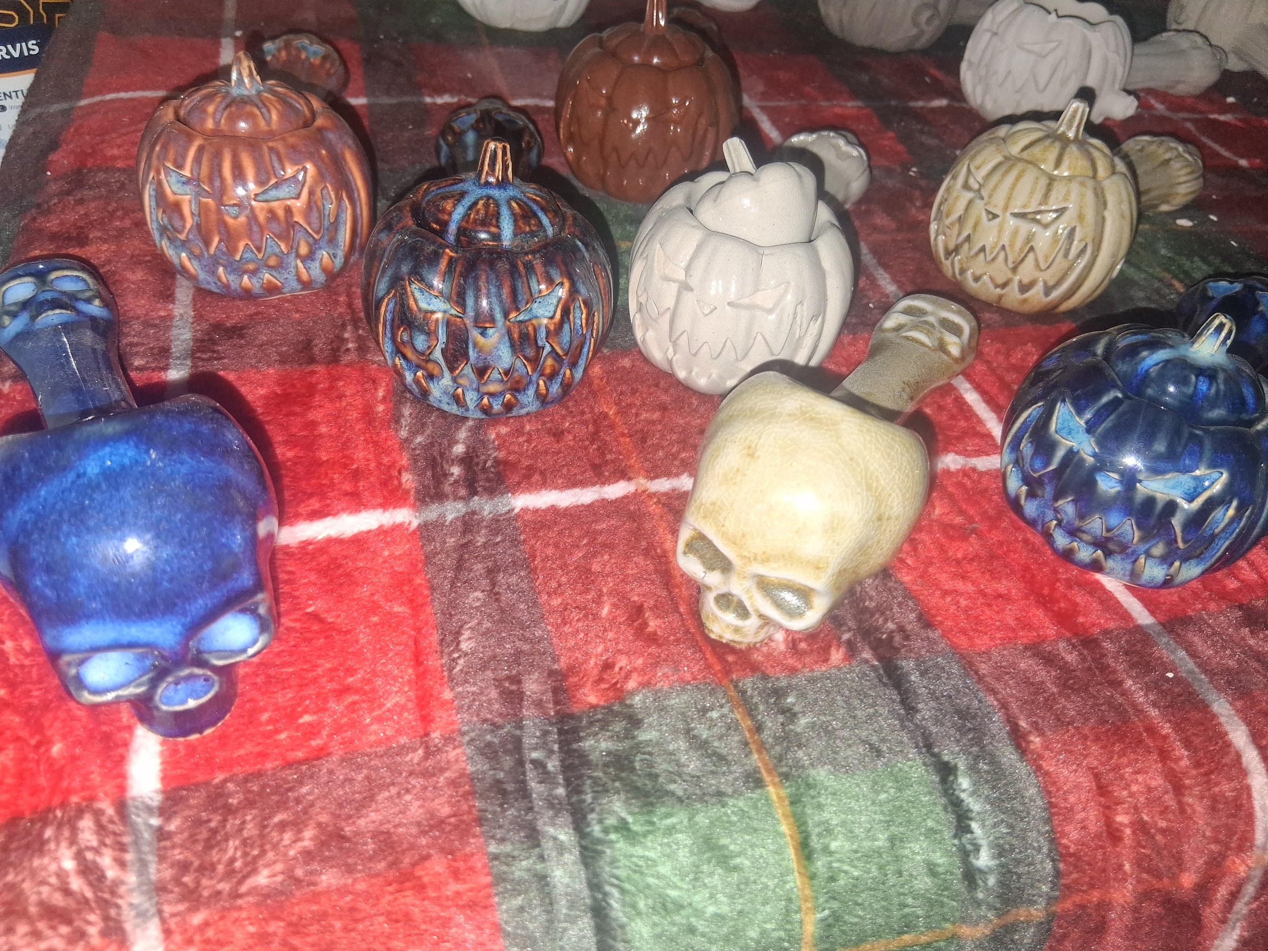 Collection of ceramic Halloween pumpkins in various colors and a ceramic skull, all arranged on a red and green plaid fabric.