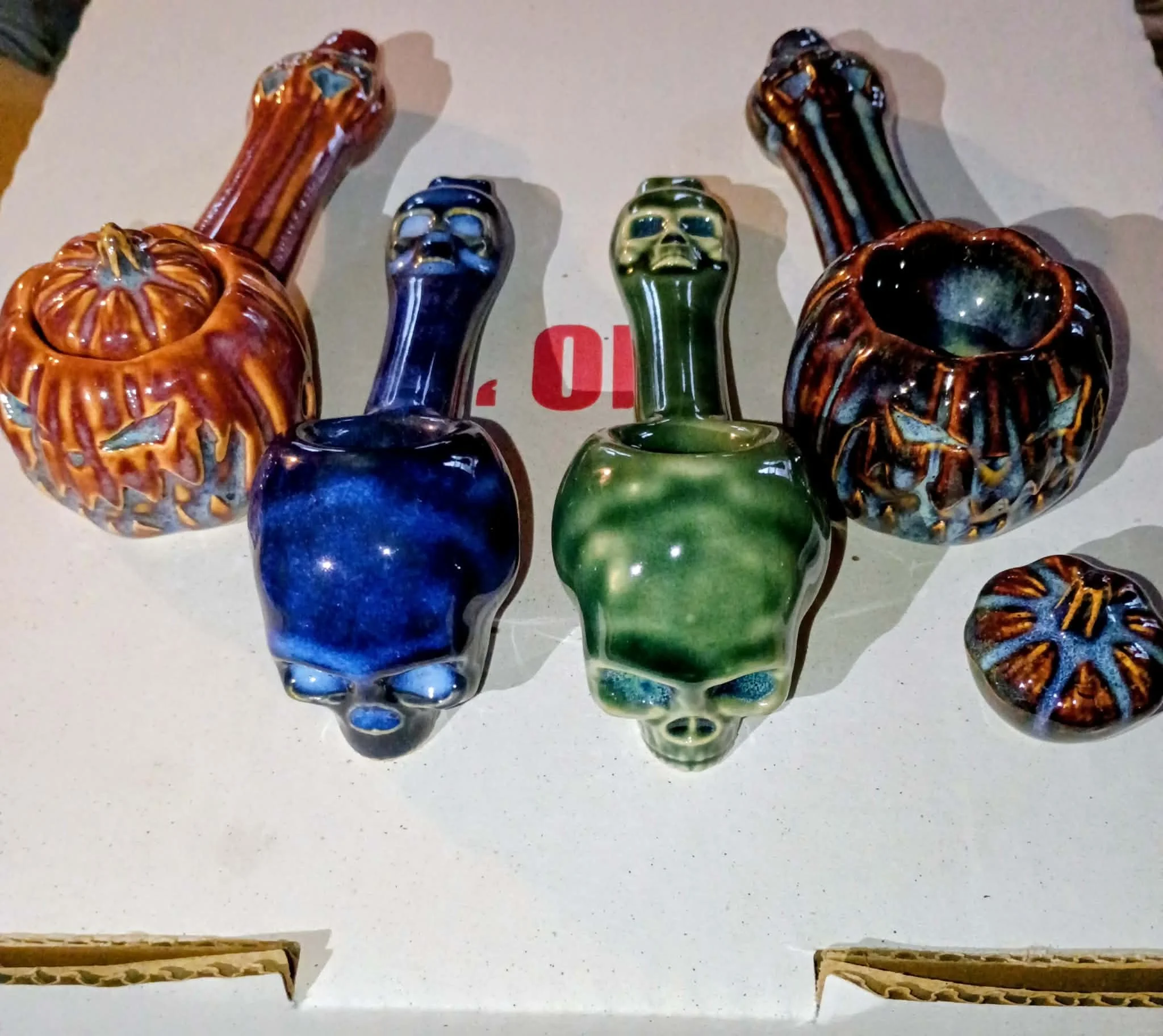 Set of ceramic pumpkin-shaped and skull-shaped pipes in orange, blue, and green glazes arranged on a white surface.