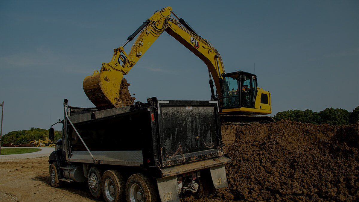 A yellow excavator loading dirt into a black dump truck on a construction site with trees and a clear sky in the background.