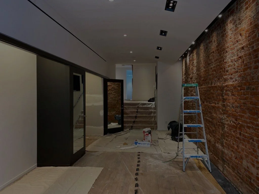 Interior space under construction with an exposed brick wall on the right, a ladder, carpeting covered with drop cloths, and construction tools and materials scattered around.