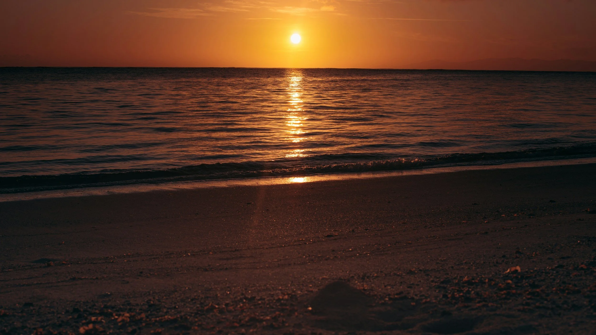 Sunset over the ocean with the sun reflecting on the water and a sandy beach in the foreground.