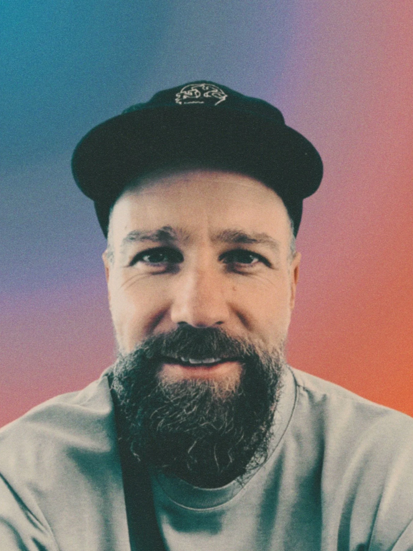 Close-up of a man with a beard wearing a black baseball cap and a light-colored shirt, smiling, with a gradient background of blue, pink, and orange.