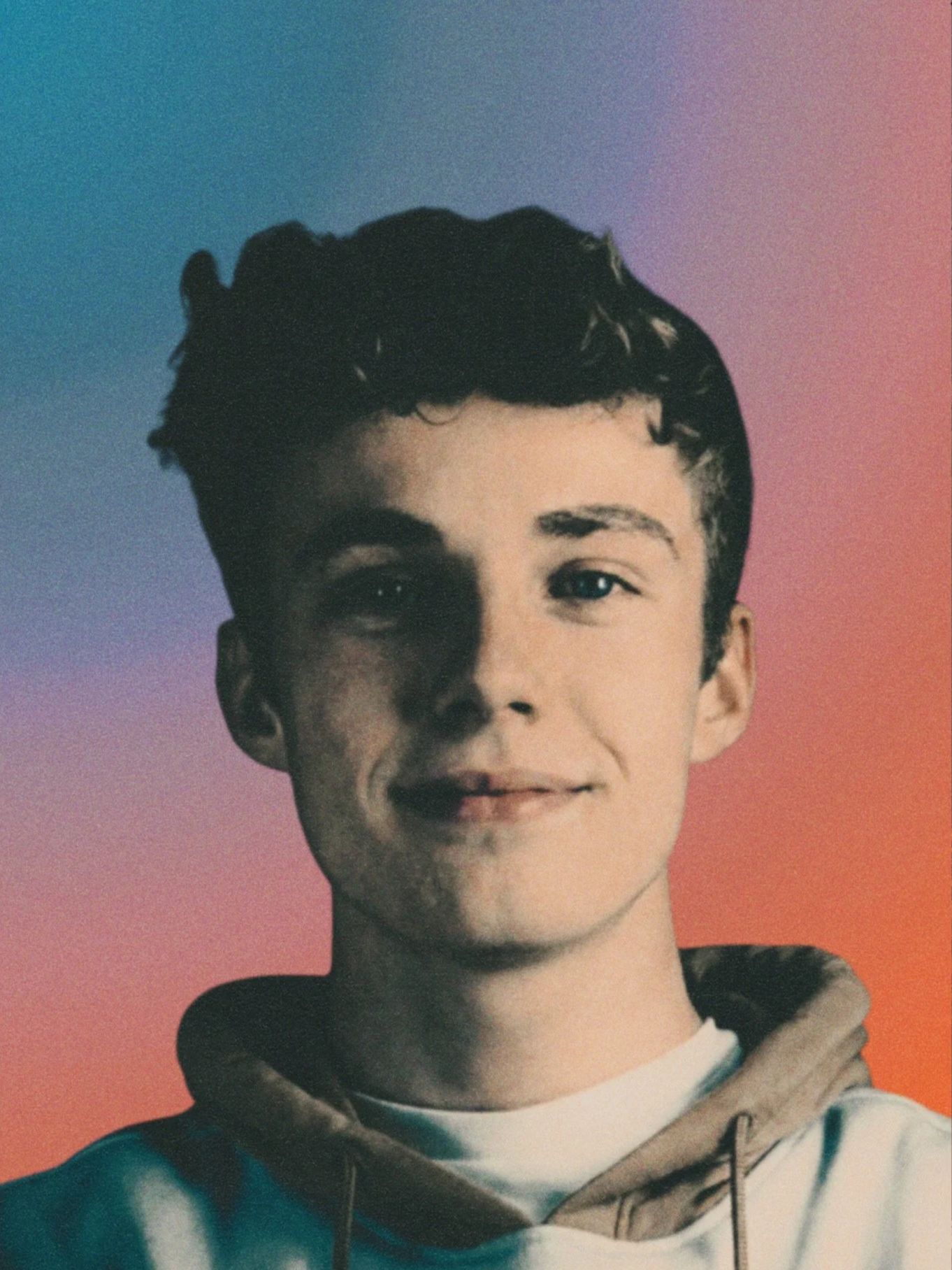 A young man with dark curly hair and a slight smile, wearing a gray hoodie, stands against a background with a gradient of blue, purple, pink, and orange colors.