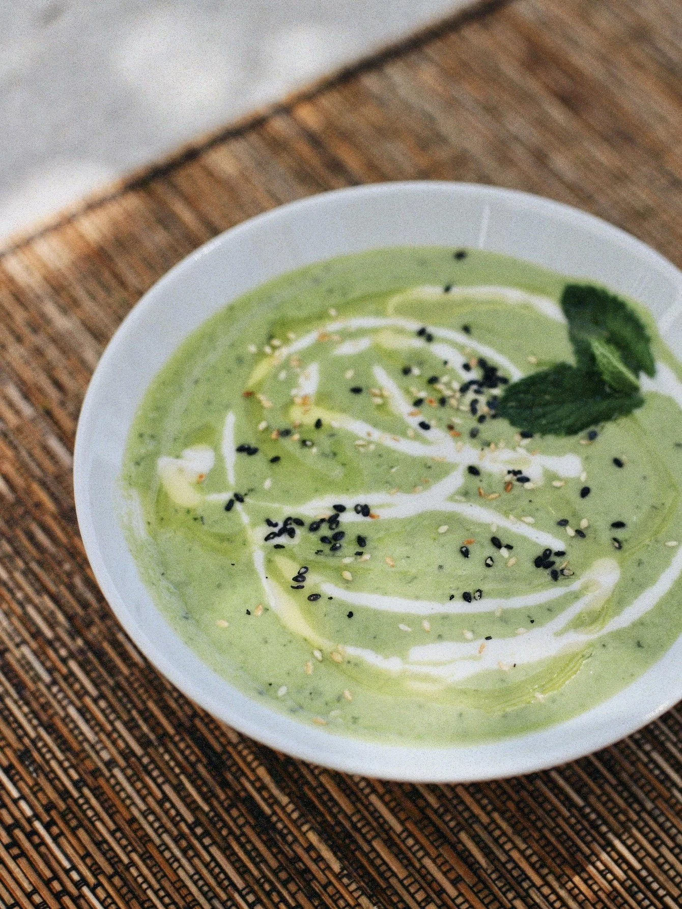 Bowl of green soup garnished with white drizzle, black sesame seeds, mint leaves, and sesame seeds, placed on a woven mat.