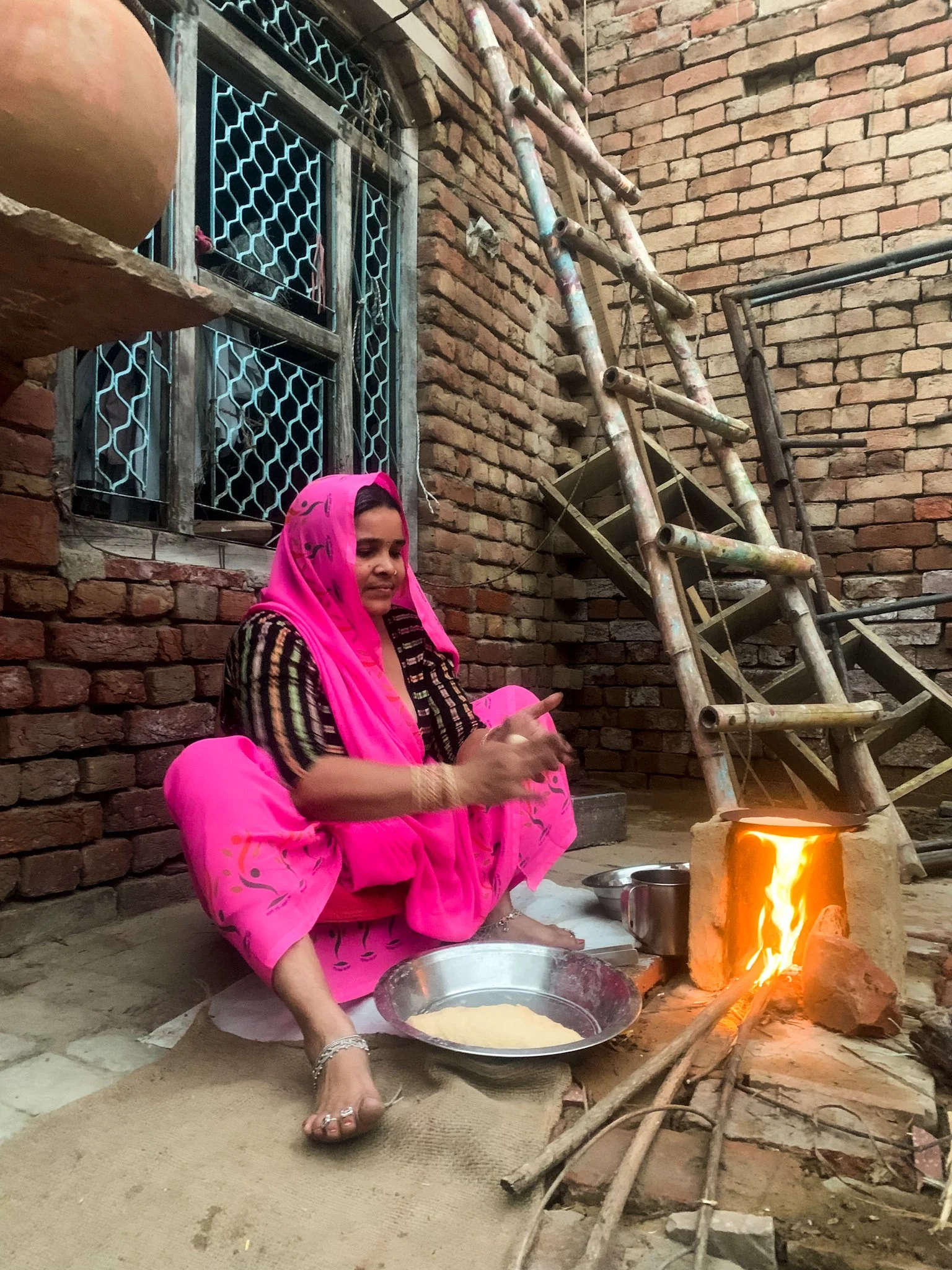 A woman dressed in pink traditional attire is sitting on the ground near a small brick stove with a fire burning inside. India Vrindavan yogi yoga bhakti himalaya retreat pilgrim rishikesh