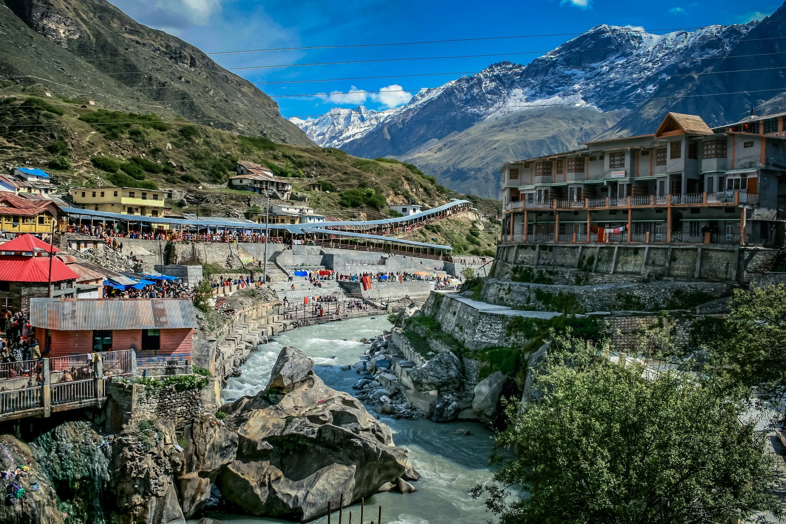Mountain village with buildings on steep hills, a river flowing through, and snow-capped peaks in the background. Vrindavan yogi rishikesh ohmat travel himalaya debbie liladhar yoga bhakti india
