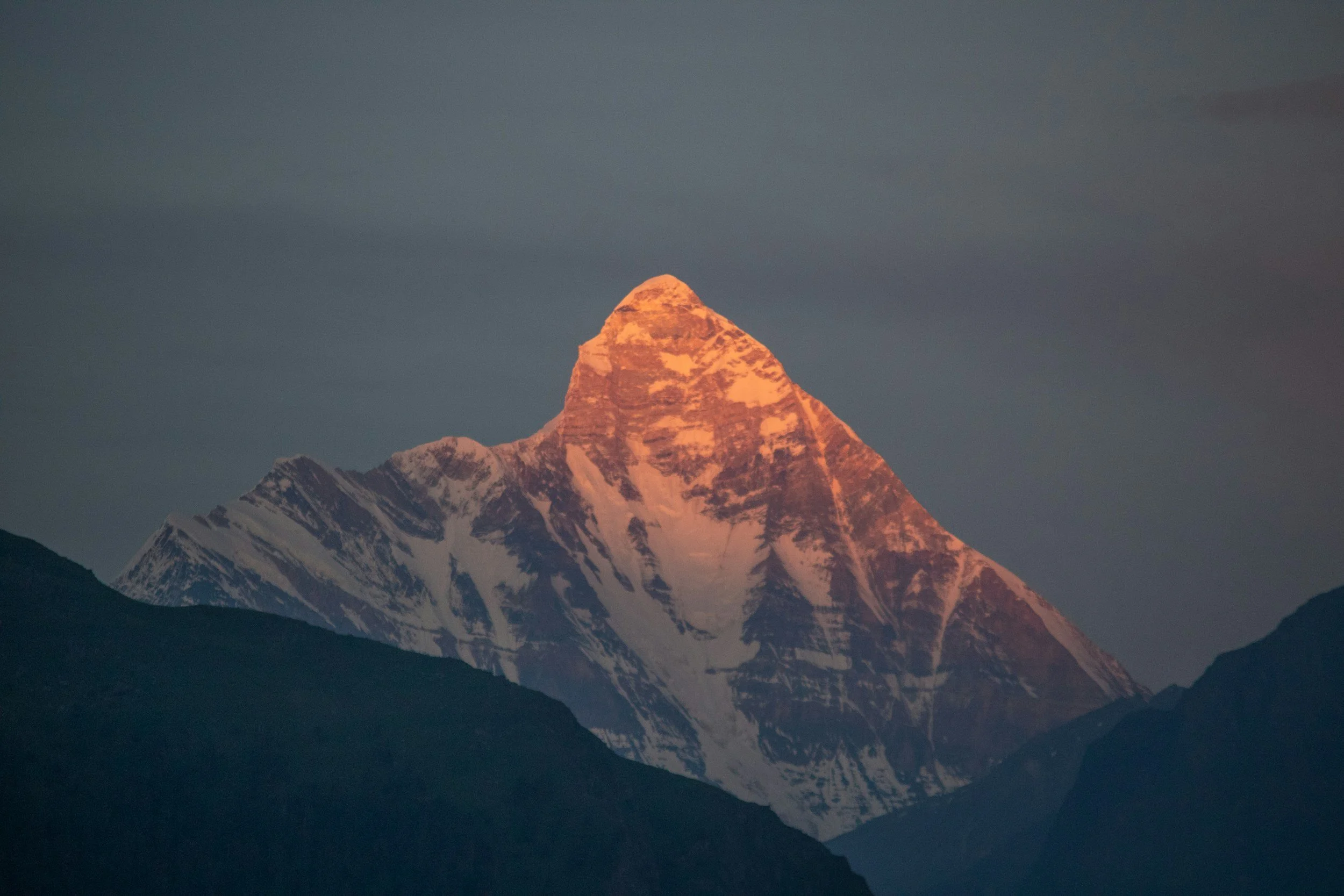 A snow-capped mountain peak illuminated by sunlight, with dark mountain silhouettes in the foreground and a cloudy sky above. Vrindavan yogi rishikesh ohmat travel himalaya debbie liladhar yoga bhakti india