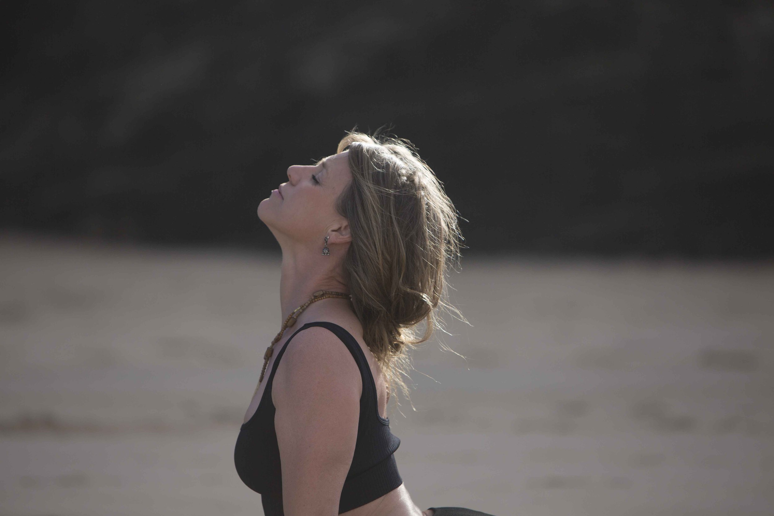 A woman practicing yoga outdoors on a beach, standing with her eyes closed and head tilted back.