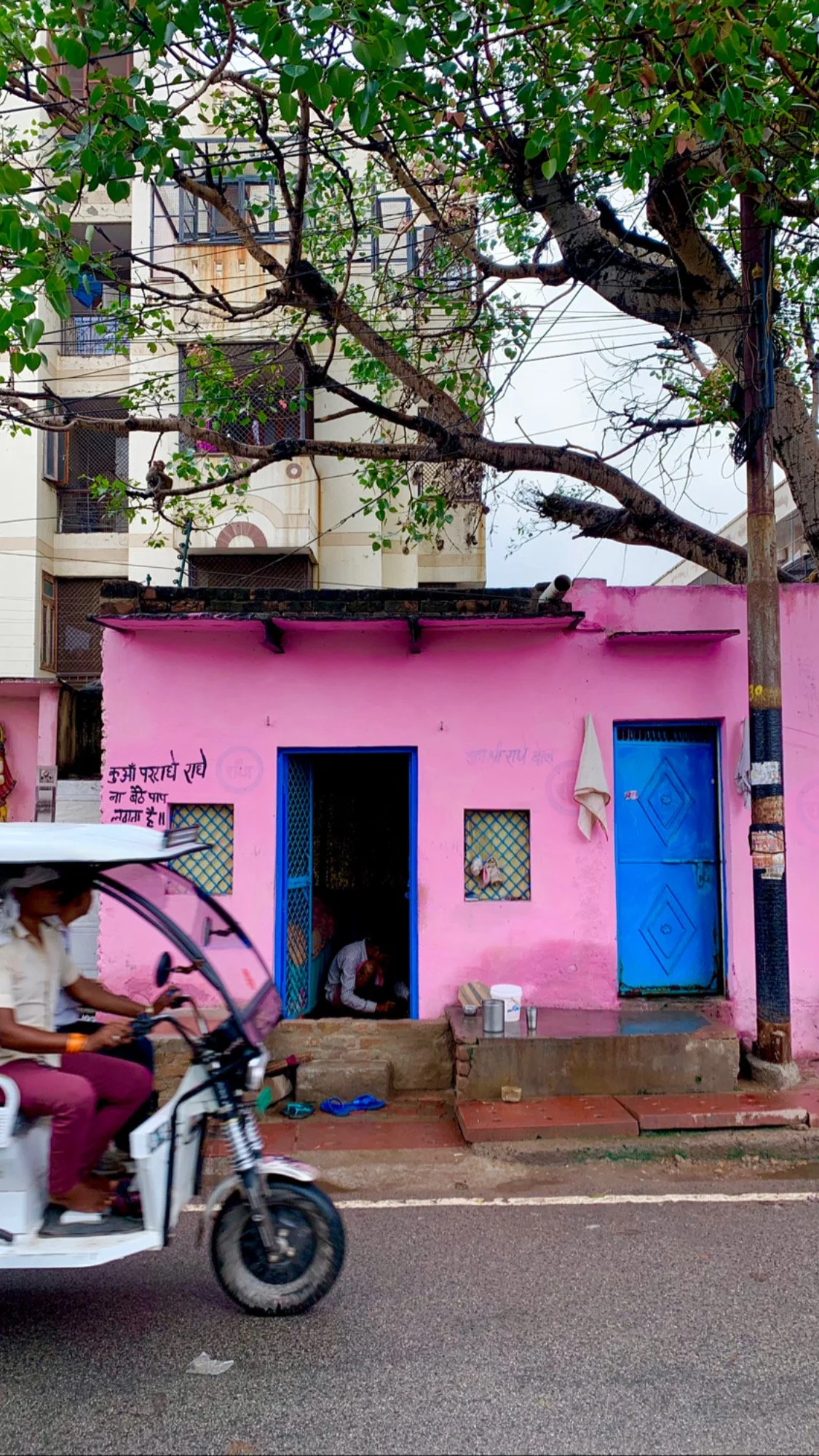 A pink building with a blue door and small barred windows, with a woman sitting outside and a person on a motorcycle passing by.