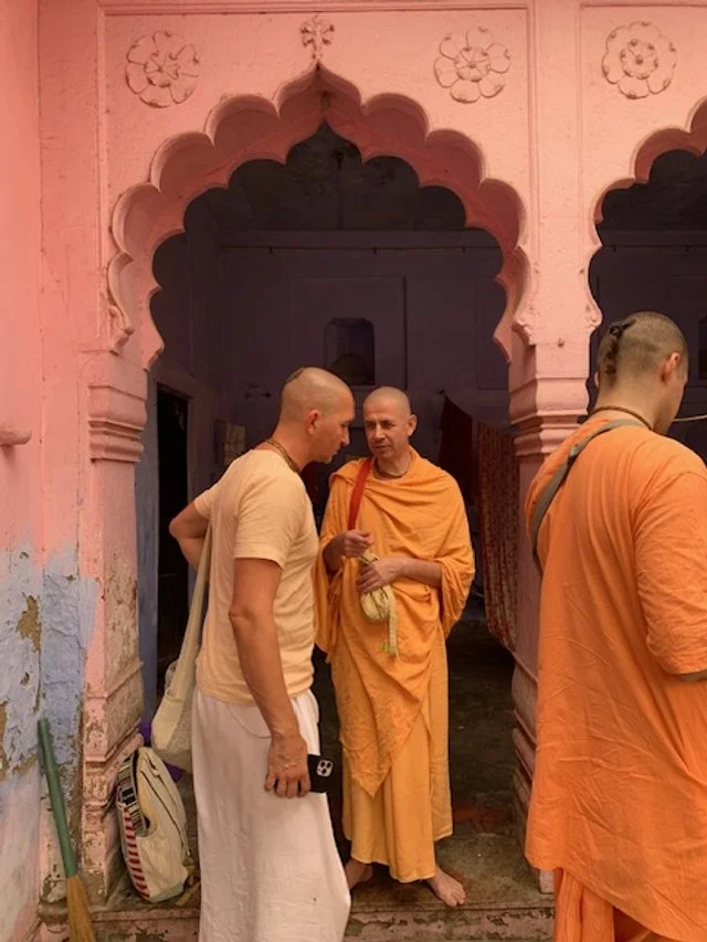 Two men in traditional Indian attire and a monk in orange robes standing under a pink stone archway with floral carvings, engaging in conversation.