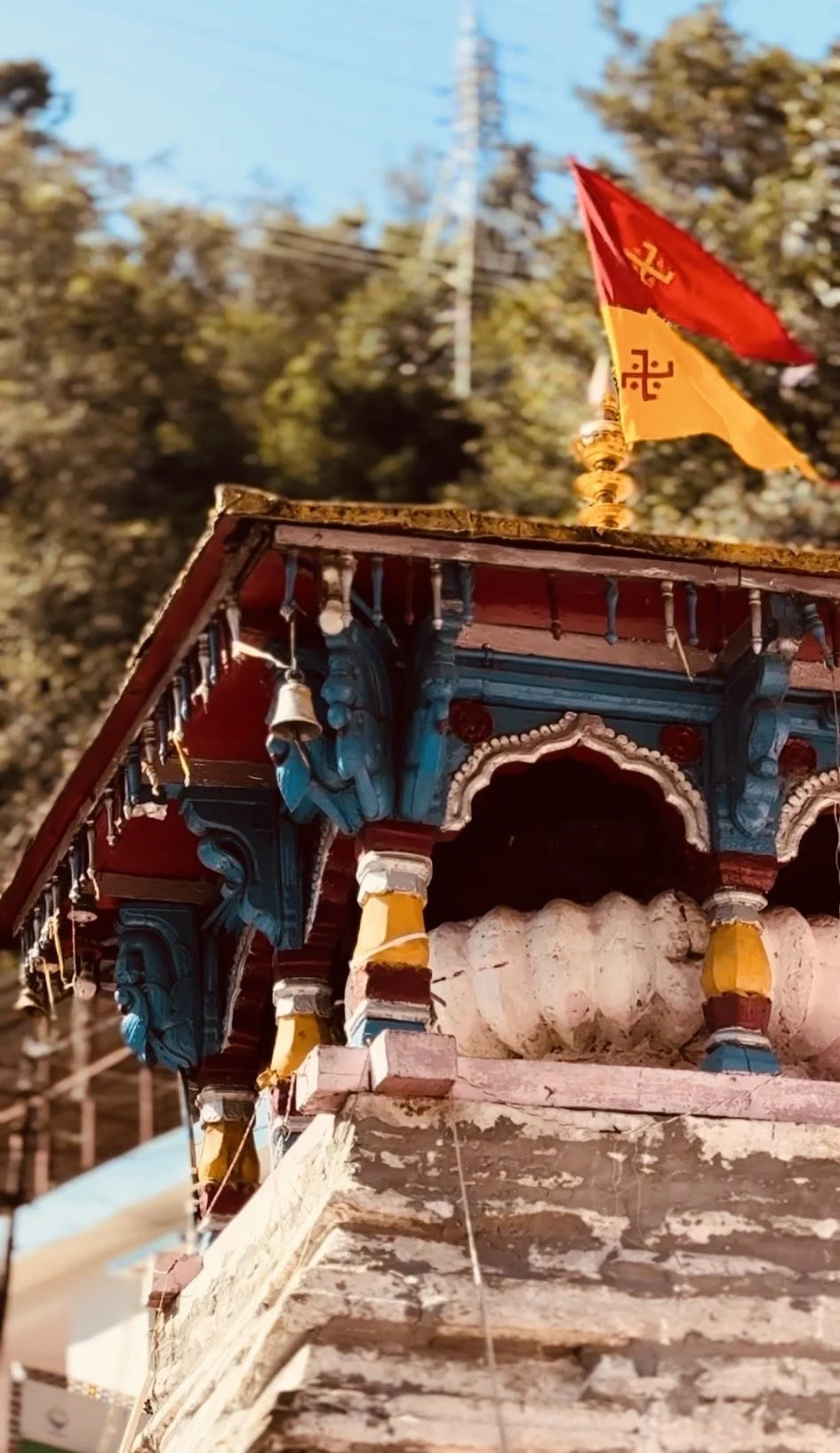 Close-up of a small temple or shrine with detailed wooden architecture, colorful paint, bells hanging from the roof, and a flag with a yellow and red design flying above. The background shows trees and a clear sky.