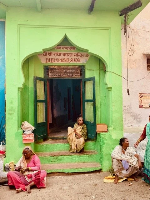 Scene outside a Hindu temple with green walls and open black doors, with four women sitting on the steps and sidewalk, one in a pink sari, one in a cream sari, another in a white sari, and one in traditional attire. A sign in Hindi is above the entra