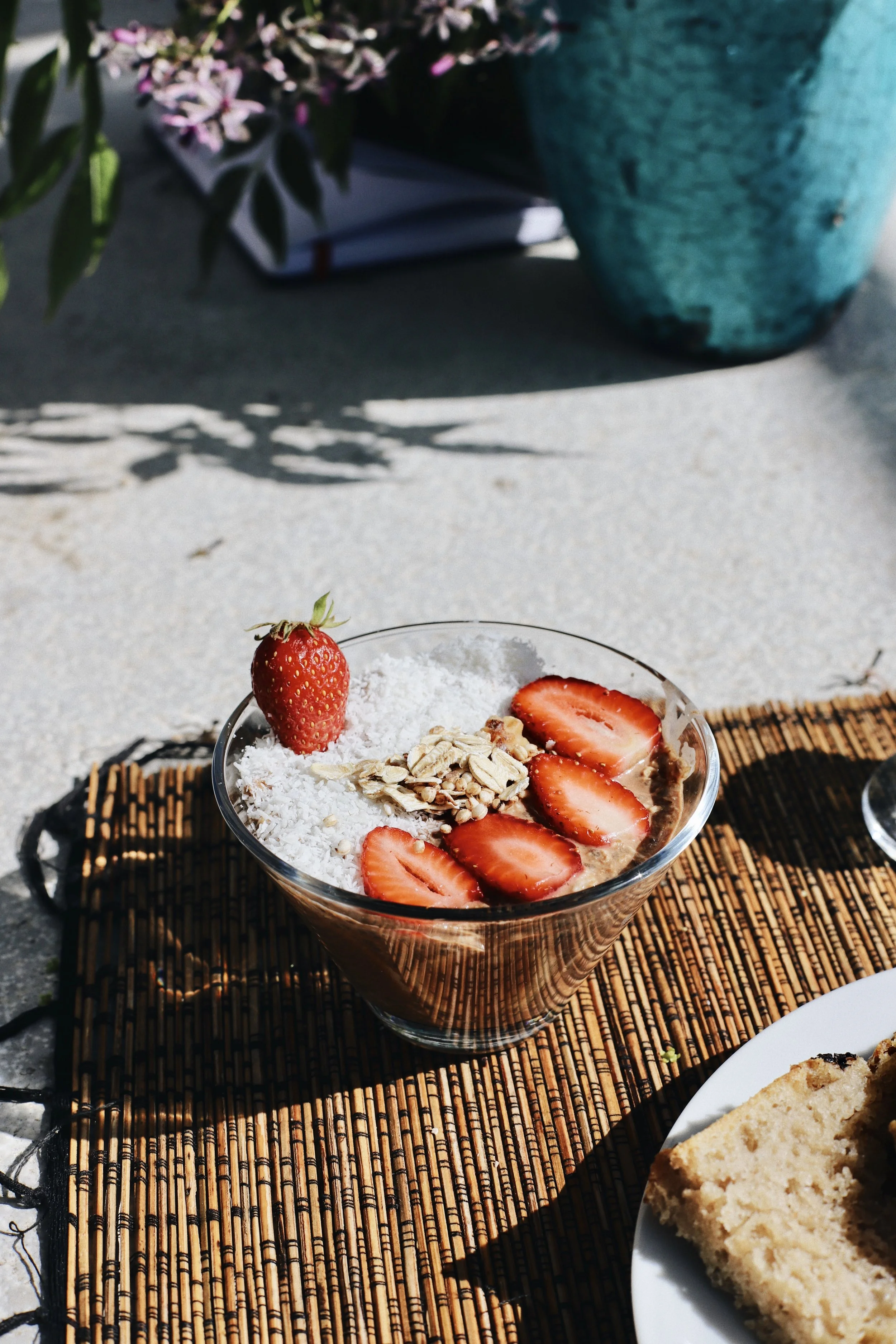 A glass bowl of chocolate mousse topped with sliced strawberries, a whole strawberry, shredded coconut, and granola, placed on a bamboo mat outdoors with sunlight.