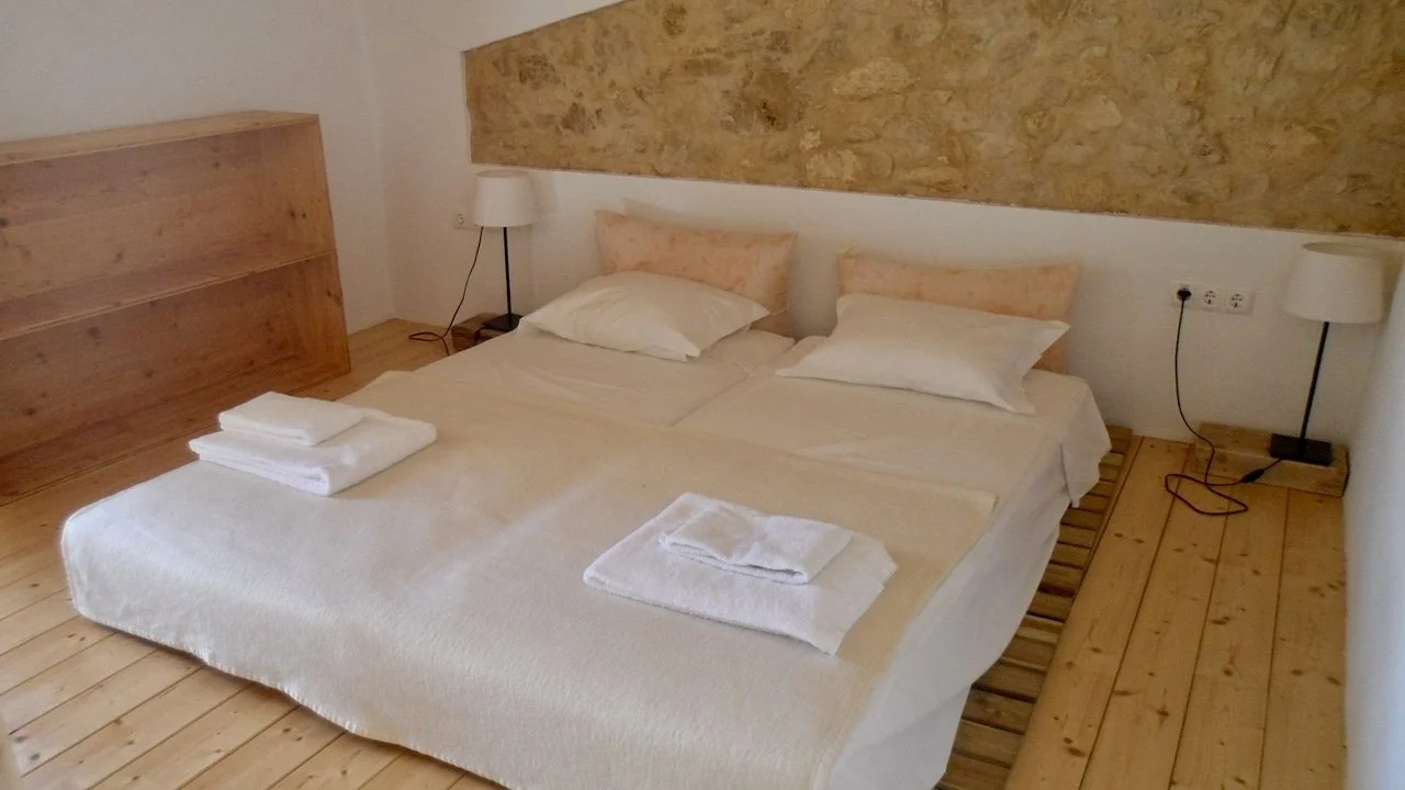 A bedroom with a double bed, two pillows, white sheets, and folded towels on the bed. There are two wall-mounted lamps on either side of the bed and wooden flooring.