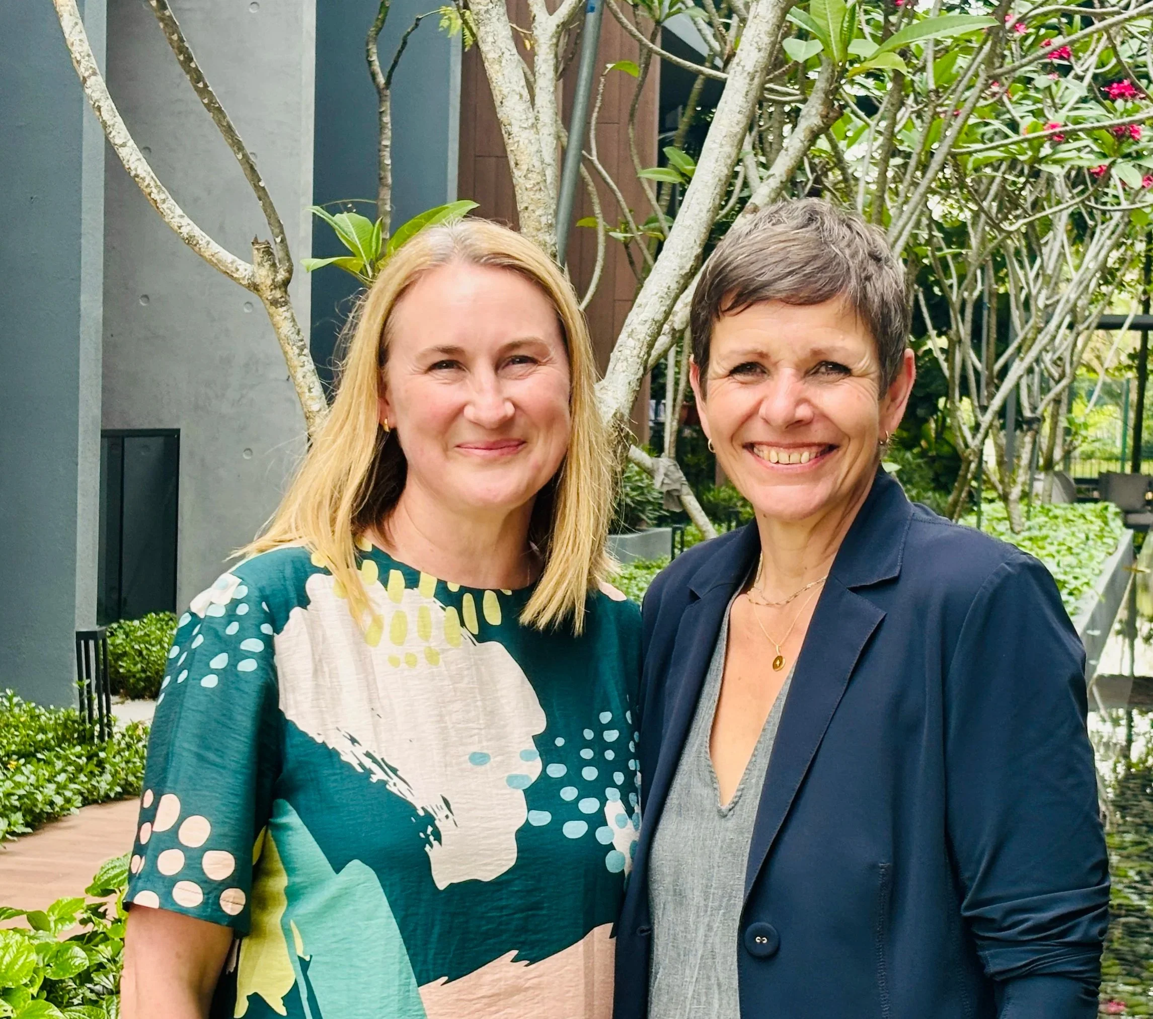 Two women smiling outdoors in front of a tree and greenery, one wearing a patterned blouse and the other in a dark blazer.