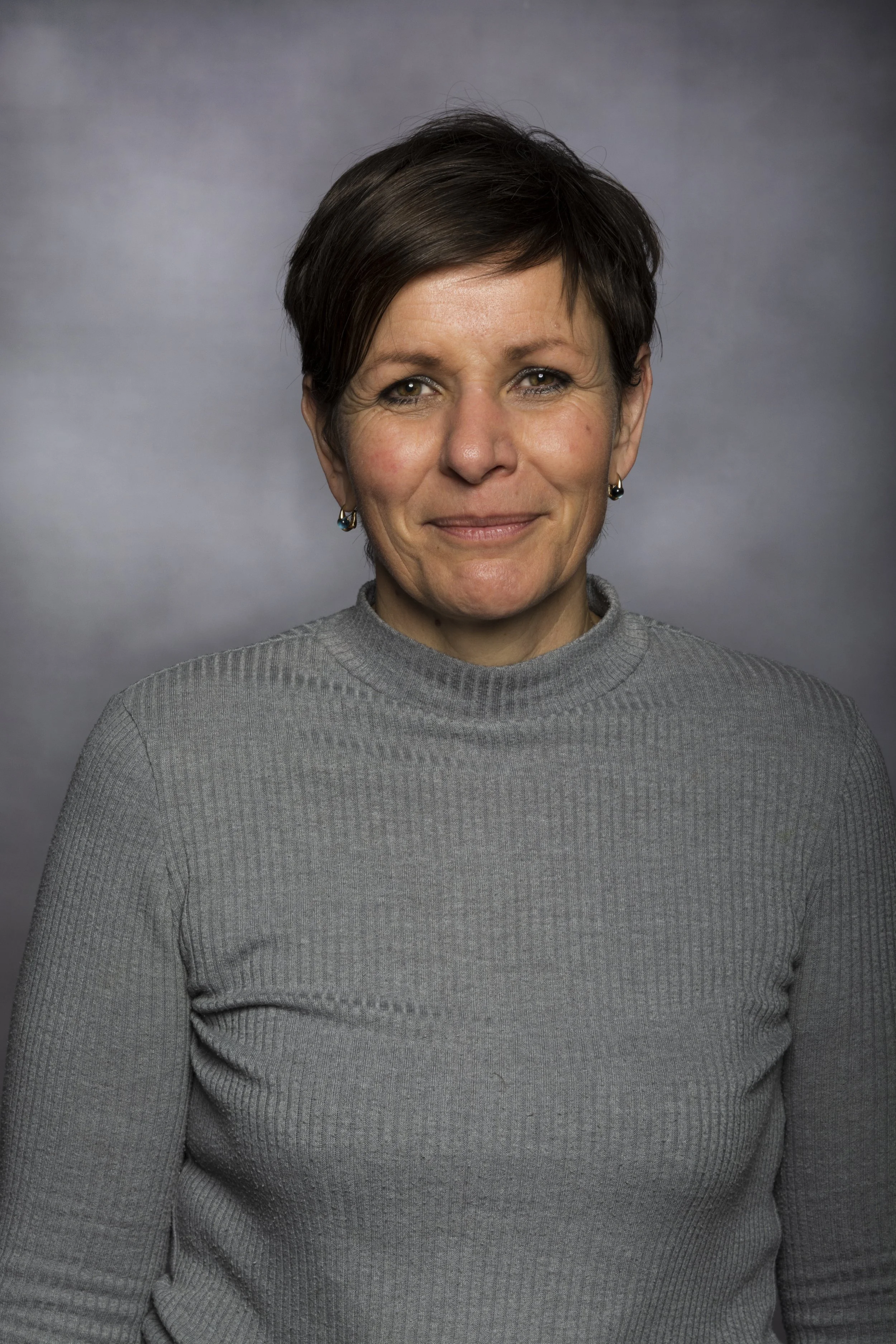 A woman with short dark hair wearing a gray ribbed turtleneck sweater and earrings, smiling slightly against a gray background.