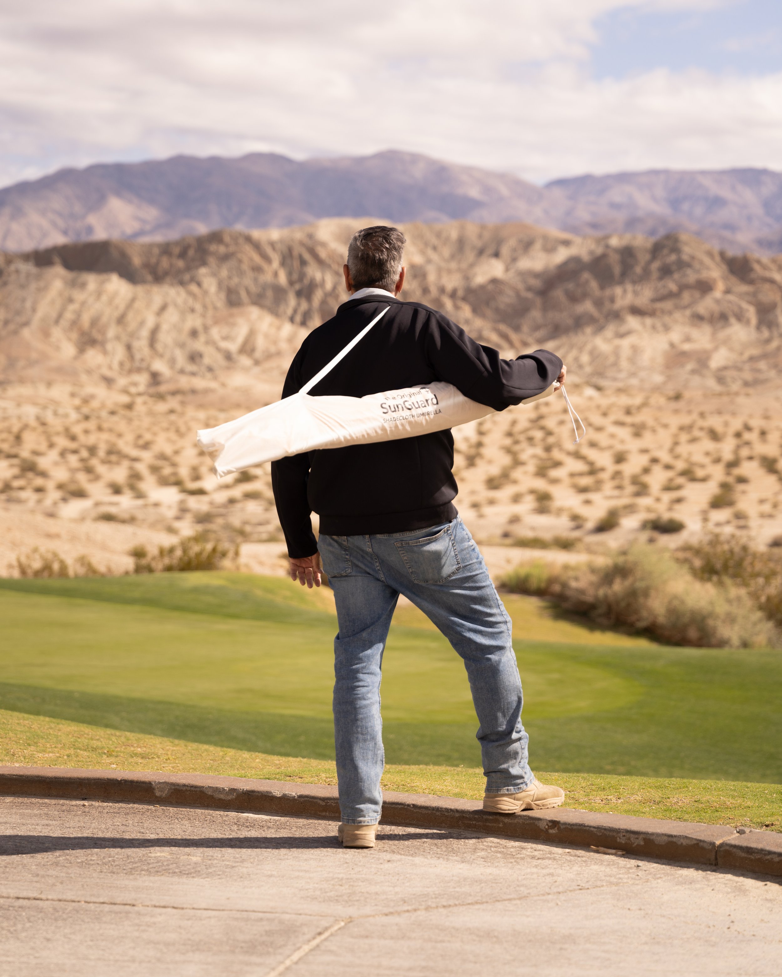 A man standing on a golf course's concrete path, holding a golf club in his right hand and a white umbrella over his shoulder, with mountainous desert landscape in the background.
