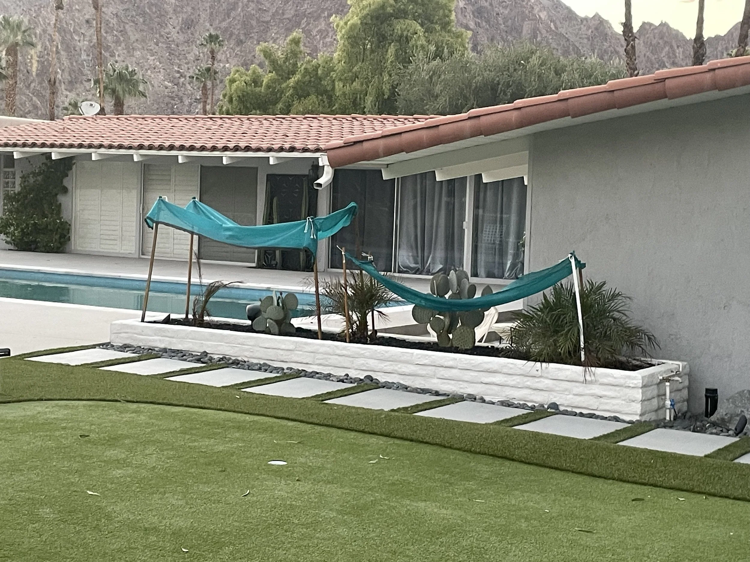 A house with a tiled roof and a backyard with a swimming pool, a blue hammock, and decorative plants, with mountains and palm trees in the background.