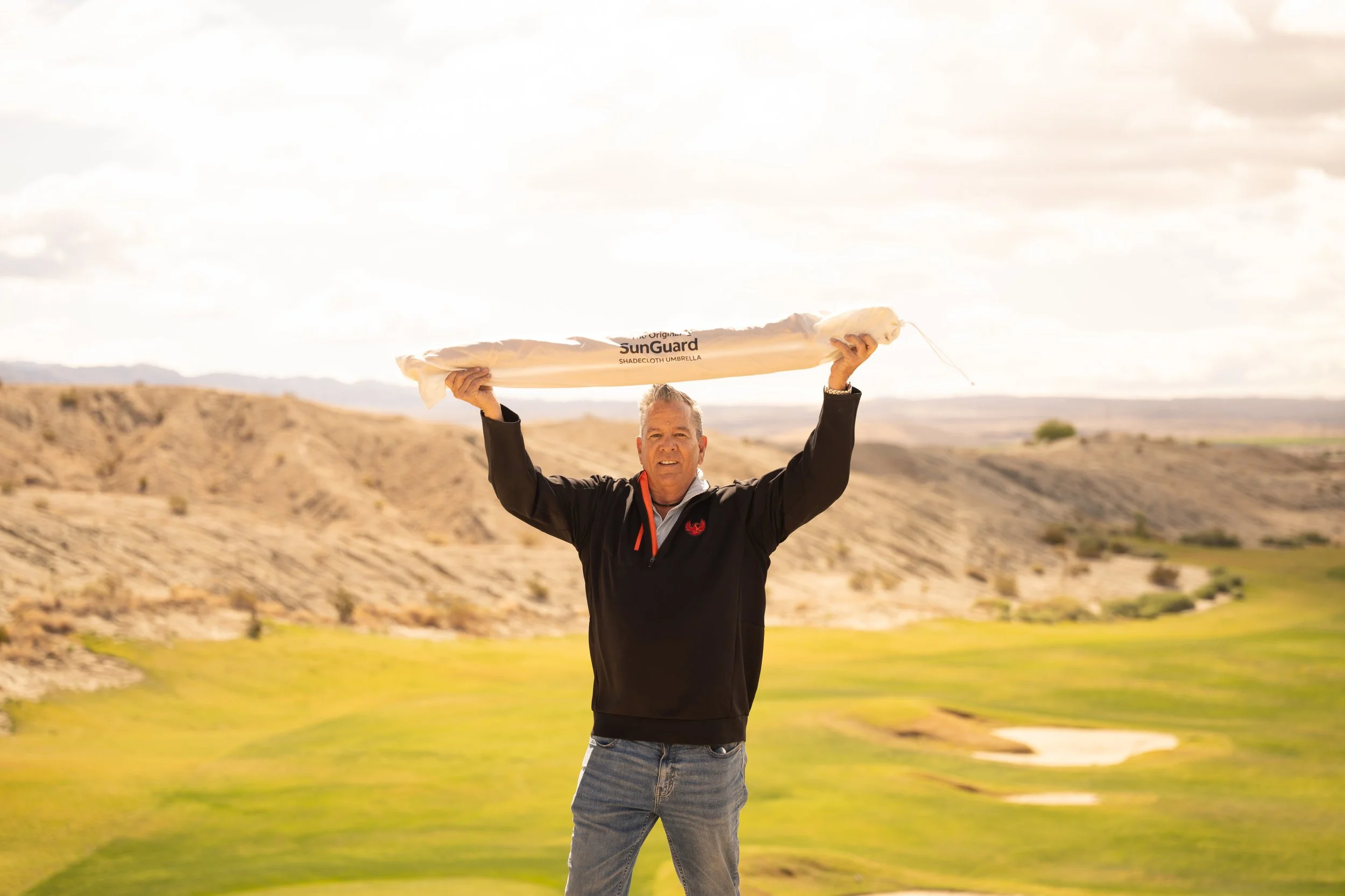 A man outdoors holding a sunshade umbrella above his head in a desert landscape with mountains in the background.