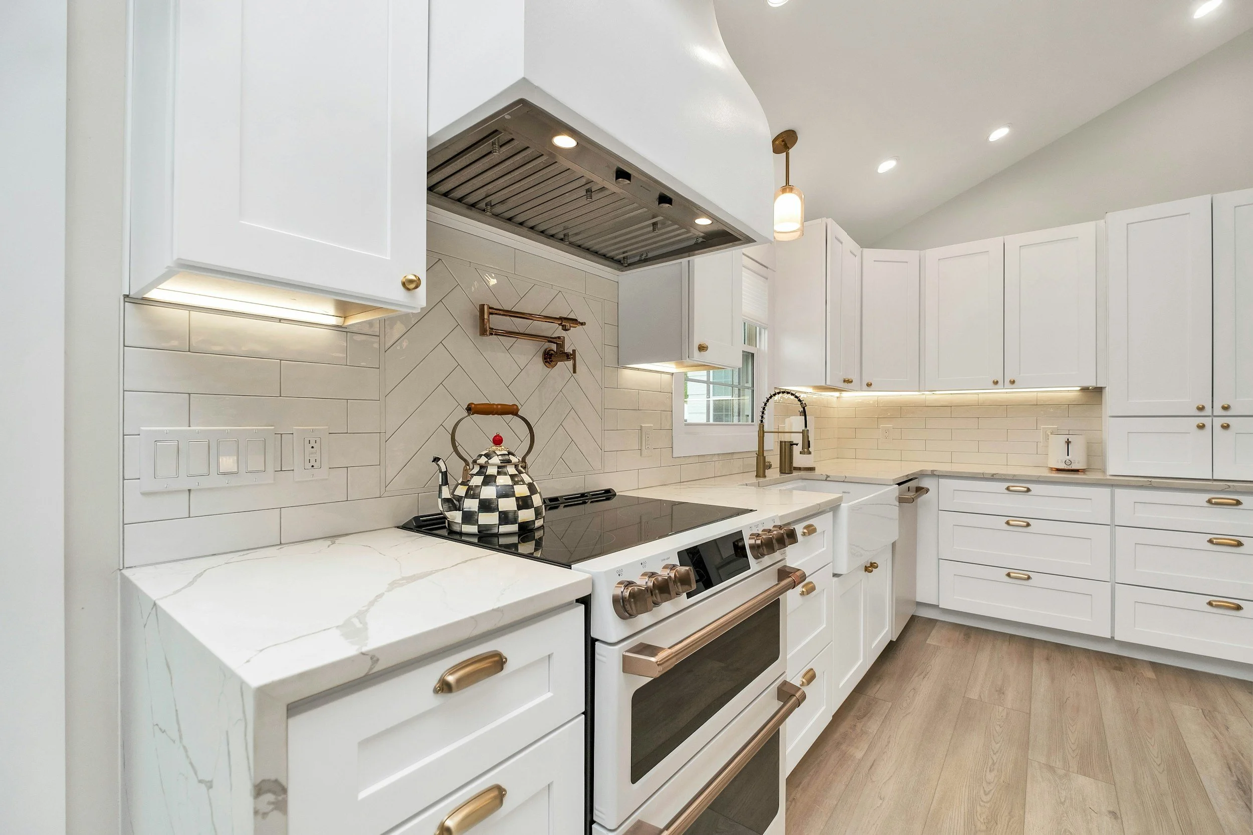 Modern white kitchen with gold hardware, marble countertops, and a checkered teapot on the stove.