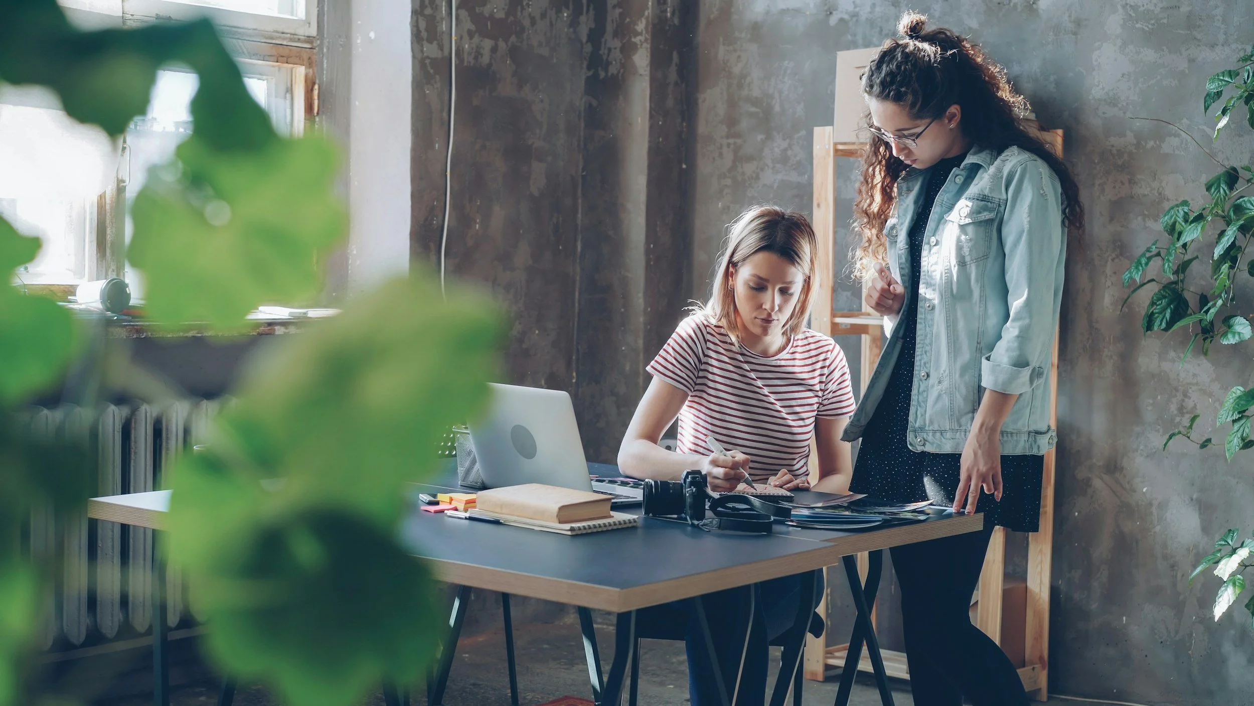 Two women are working at a cluttered desk in an industrial-style workspace. One is seated and writing, the other standing and looking at the papers. The desk has a laptop, camera, books, and notebooks. There are green plants and a large window with natural light.