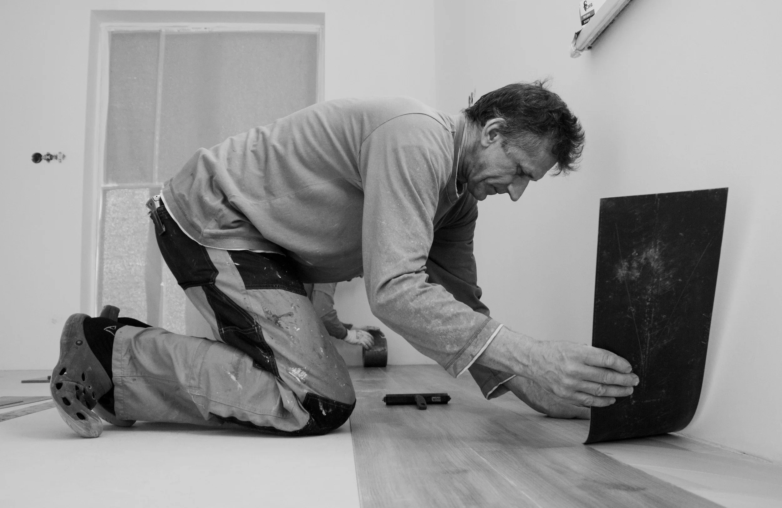 A man kneels on the floor while installing hardwood flooring, using a tool to secure the planks against the wall.