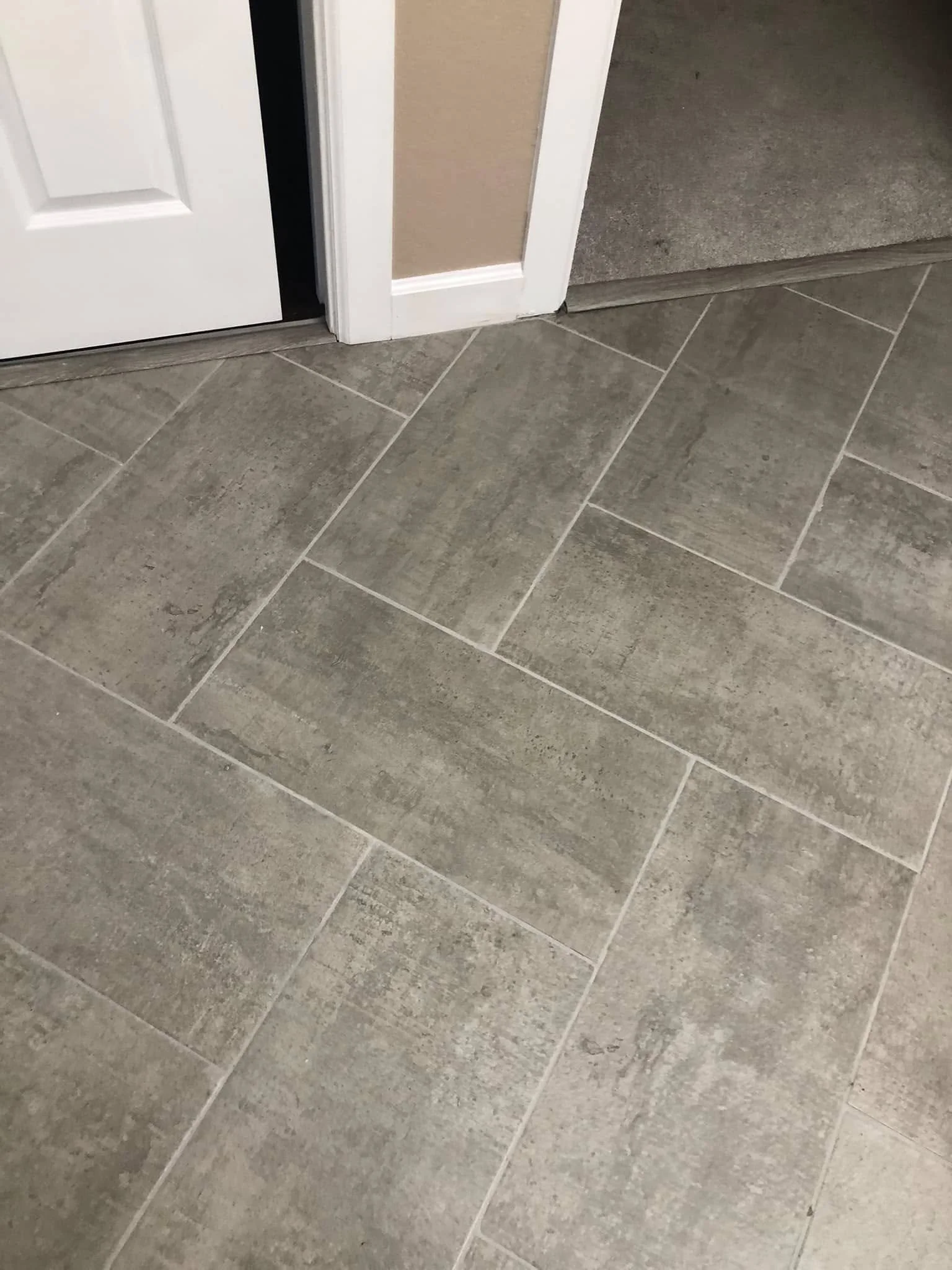 Close-up of tiled floor near a doorway, with beige and gray tiles, and a white door frame.