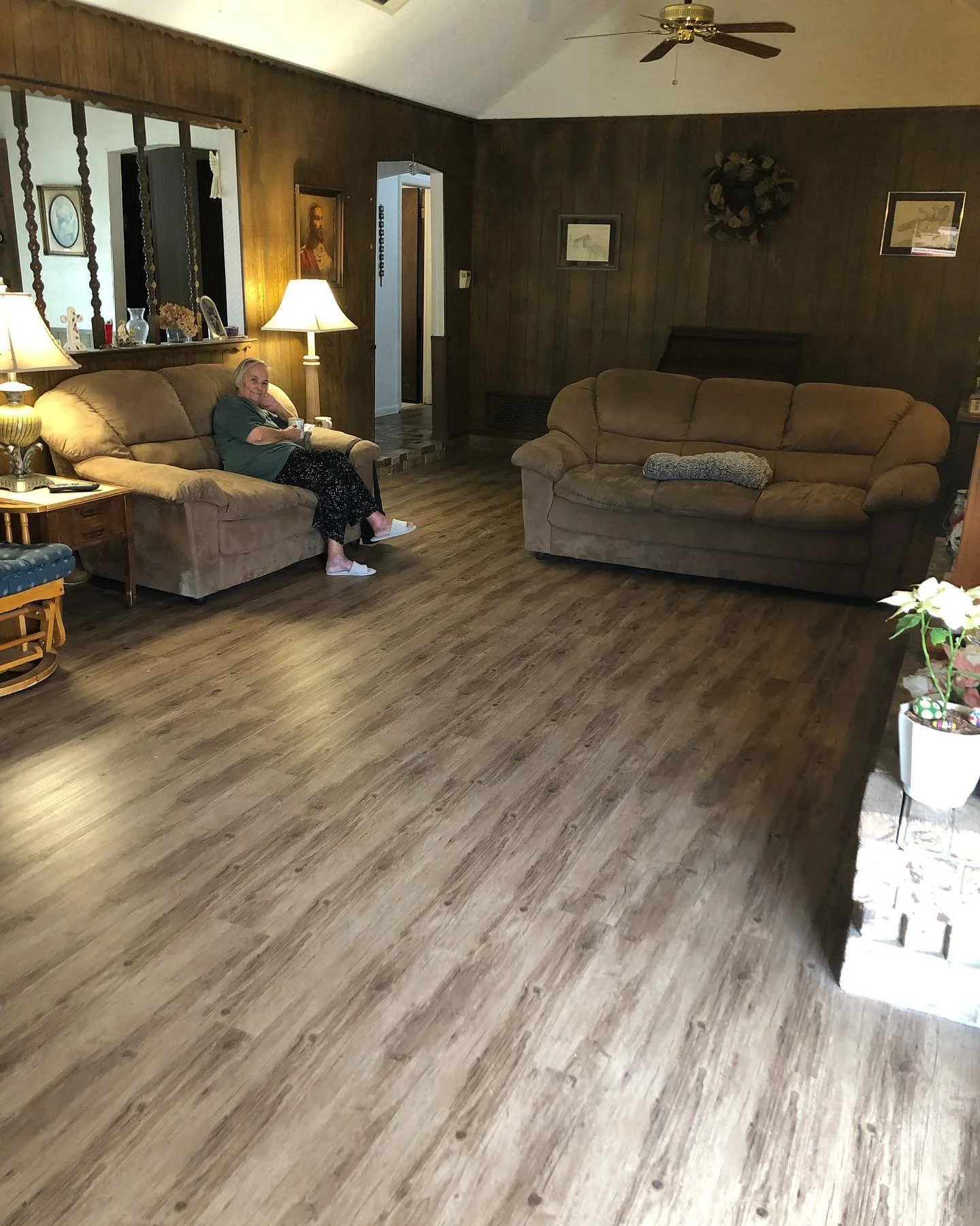 Living room with wooden walls and flooring, two beige sofas, a woman sitting on the left sofa, a table with lamps, framed pictures on the walls, and decorative items.
