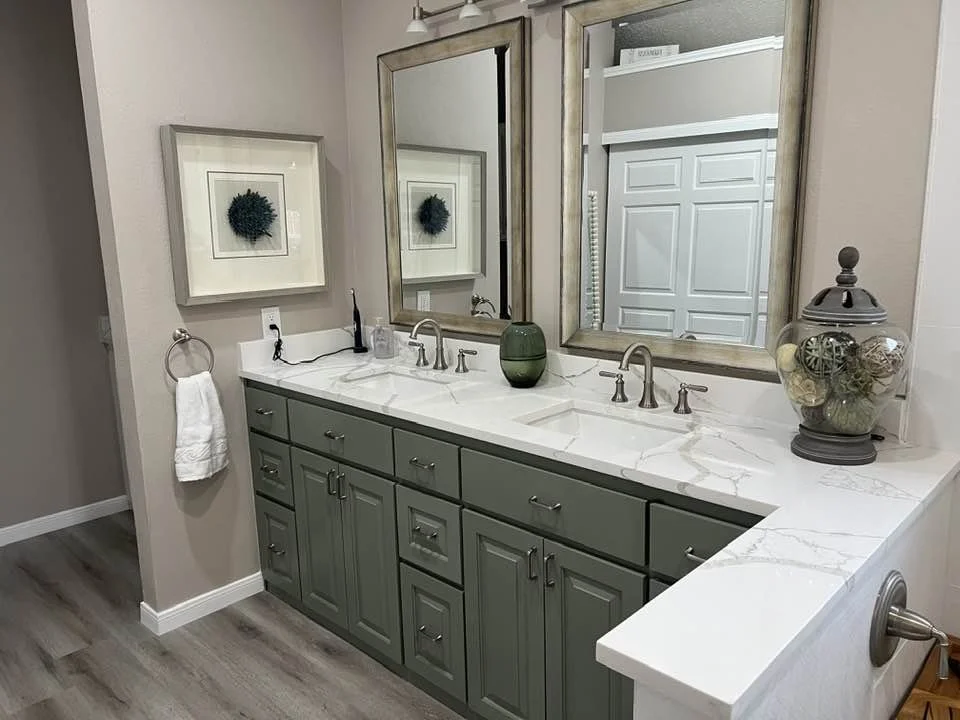 Bathroom vanity area with two mirrors, green cabinets, marble countertop, and decor including a green vase and a glass jar with decorative balls.