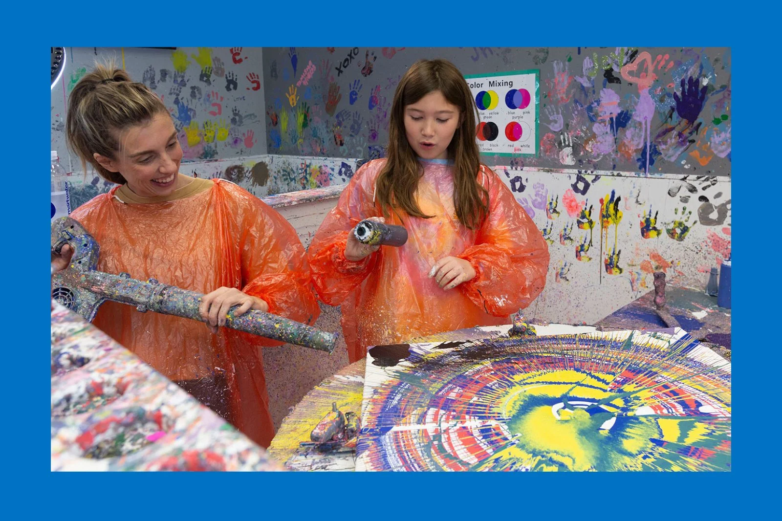 Two girls and an adult engaging in splatter painting in a colorful art studio, wearing orange protective smocks. One girl holds a paint roller, and the other has a plastic tool. The wall behind them is covered in handprints, paint splashes, and a color mixing chart. The table in front has a large circular canvas with vibrant abstract paint patterns.