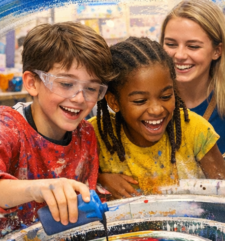 Three children joyful in art class, two girls and a boy, with paint on their clothes, using a spin art machine, and a smiling teacher in the background.