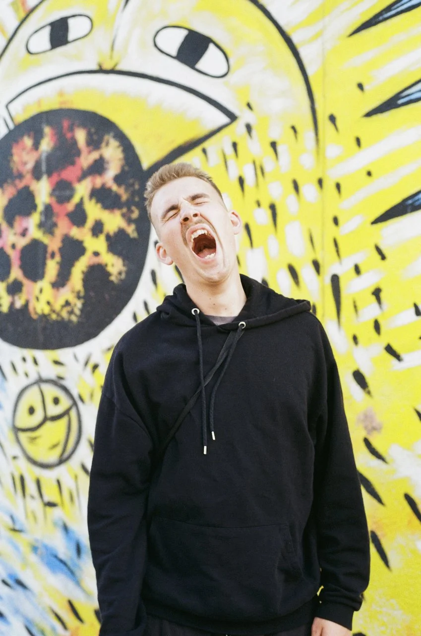 Junger Mann in schwarzer Kapuzenjacke vor bunter Wand mit lachendem und schreien Ausdruck.