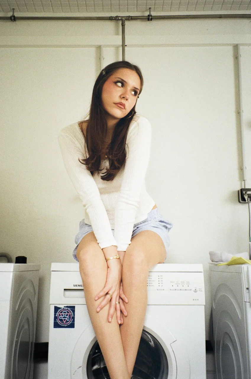 Eine junge Frau sitzt auf einer Waschmaschine in einer Waschküche. Sie trägt ein langärmeliges weißes Oberteil und kurze Shorts und blickt nach oben und zur Seite.
