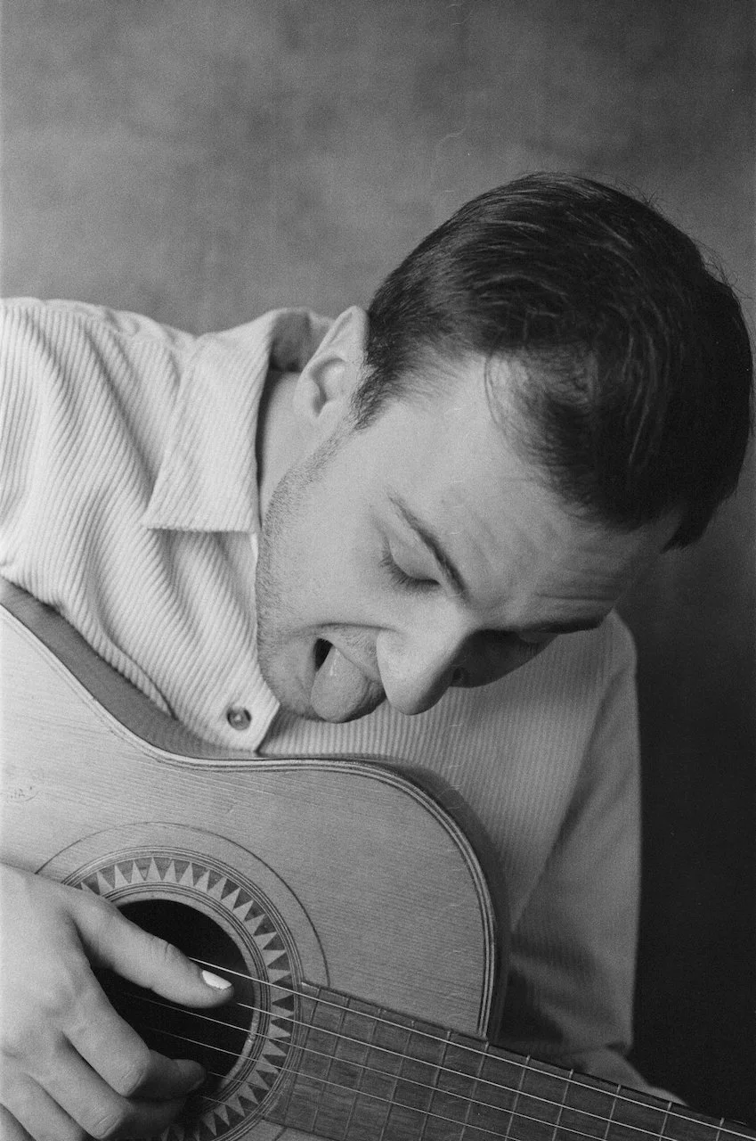 Schwarz-Weiß-Foto eines Mannes, der ein E-Gitarre spielt. Er hat kurze, dunkle Haare und trägt ein gestreiftes Hemd. Der Mann ist beim Spielen, seine Zunge ist leicht herausgestreckt.