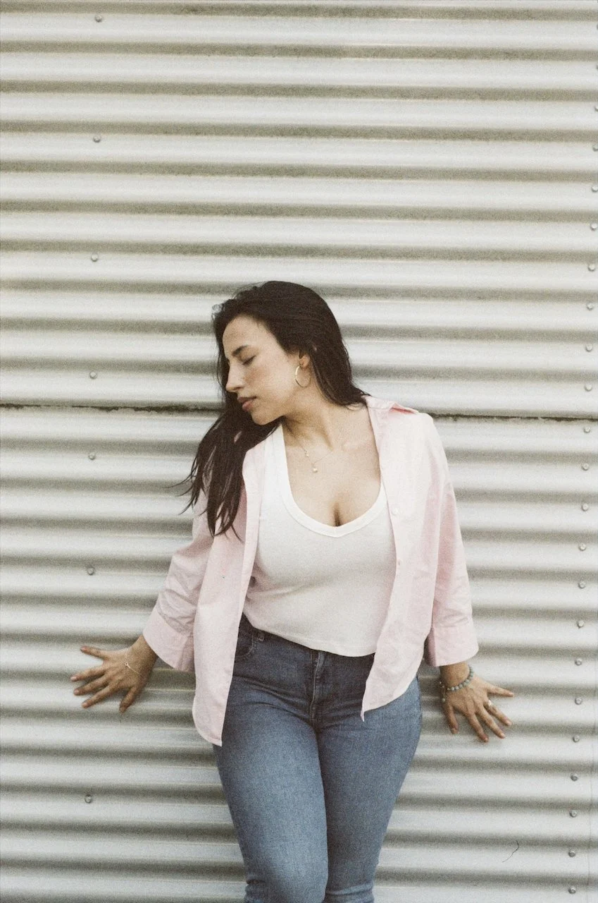 Junge Frau lehnt an einer Metallwand, trägt weiße T-Shirt und eine rosa Jacke, mit dunklen Haaren und großen Ohrringen.