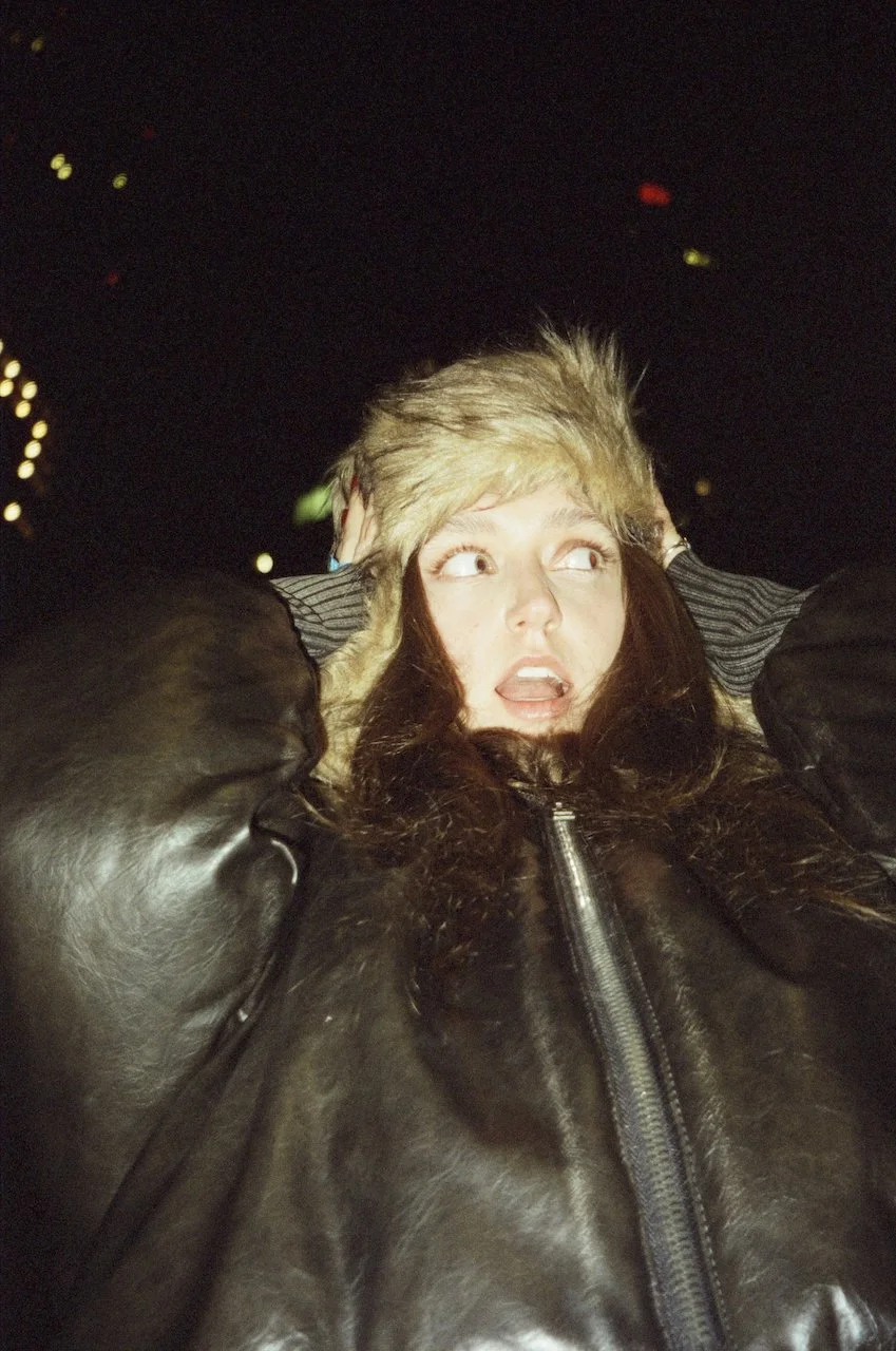 Nahaufnahme einer Frau, die überrascht oder schockiert aussieht, bei Nacht, trägt eine Pelzmütze und eine dunkle Lederjacke.