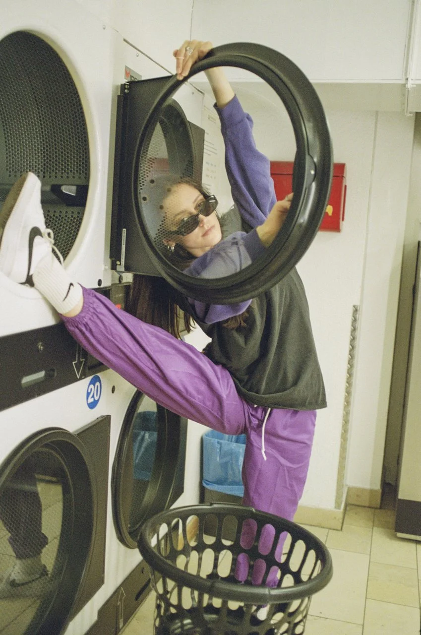 Junge Frau im Waschsalon mit Sonnenbrille, die eine Wäsche in die Waschmaschine steckt.