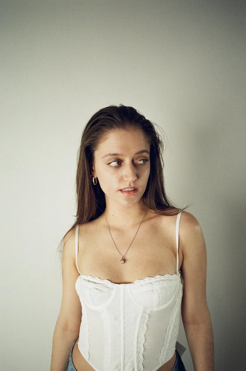 Junge Frau mit braunen Haaren trägt ein weißes Korsett und einen dünnen Goldkettchen mit Anhänger, steht vor einer hellen Wand, schaut seitlich.