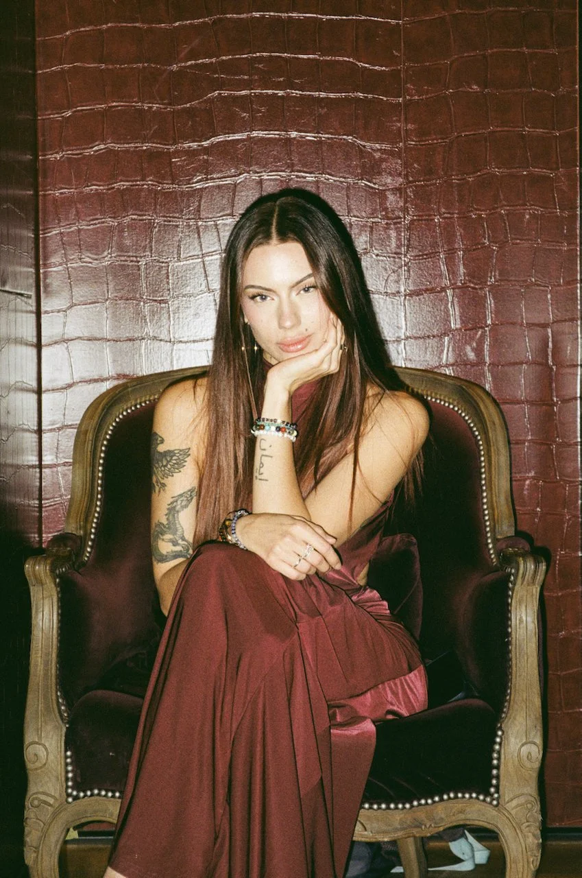 Eine junge Frau sitzt auf einem antiken, dunkelgrünen Sessel vor einer roten, texturierten Wand. Sie hat lange braune Haare, trägt einen bordeauxfarbenen Kleiderschrank und mehrere Armbänder sowie Ringe. Ihre rechte Hand liegt an ihrem Kinn, während 