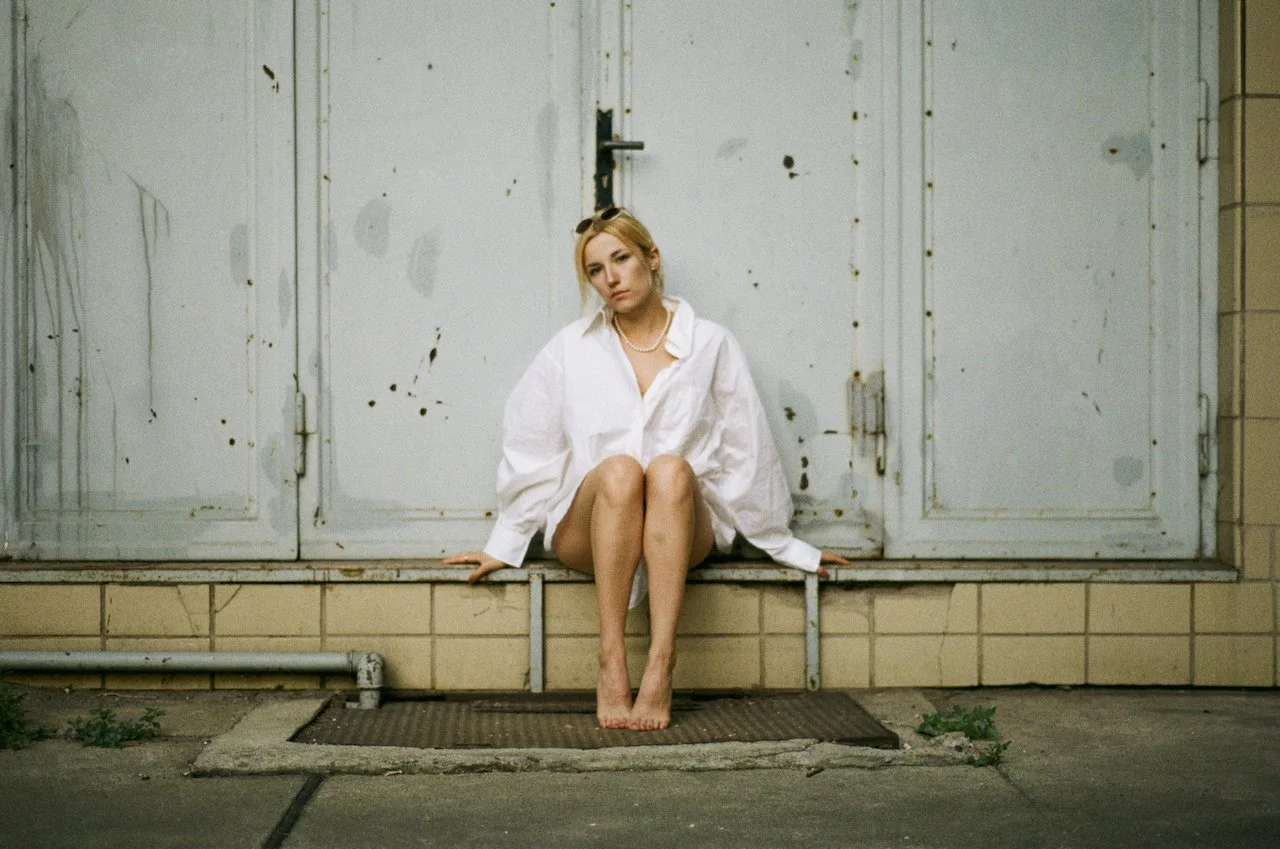 Junge Frau sitzt barfuß auf einem Bordstein vor einer alten, grauen, verschmutzten Tür, trägt ein zu groß geratenes weißes Hemd und schaut nachdenklich.