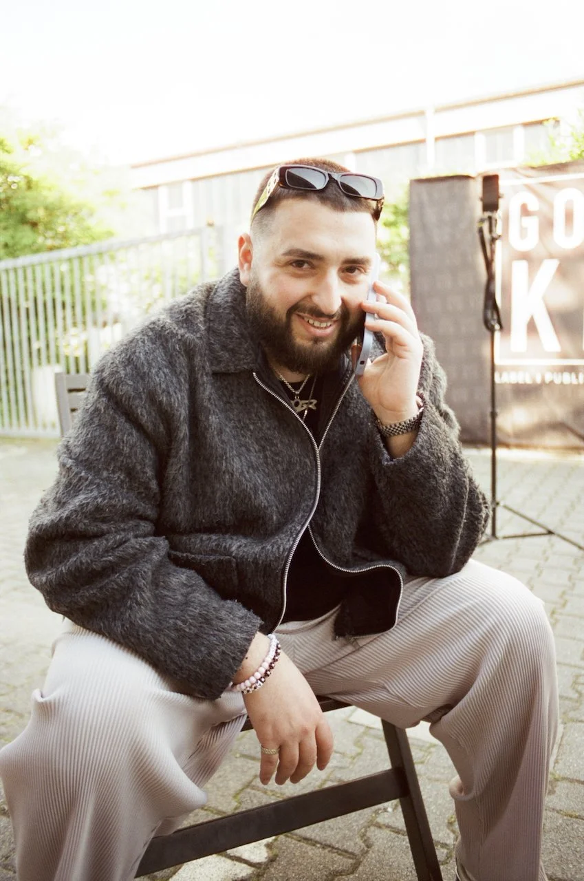 Ein lächelnder Mann sitzt auf einem Stuhl im Freien, trägt eine dunkle Jacke und beige Hose, hat Sonnenbrille auf dem Kopf und spricht am Telefon.