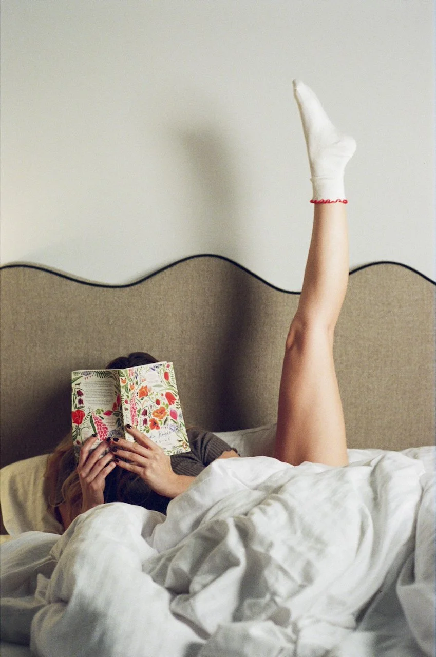Eine Person liegt im Bett, liest ein Buch mit buntem, floralem Umschlag, während sie eine Hand in die Höhe streckt. Das Bett hat weiße Bettwäsche, und die Person trägt einen grauen Pullover. Im Hintergrund befindet sich eine beige Kopfleiste an der W