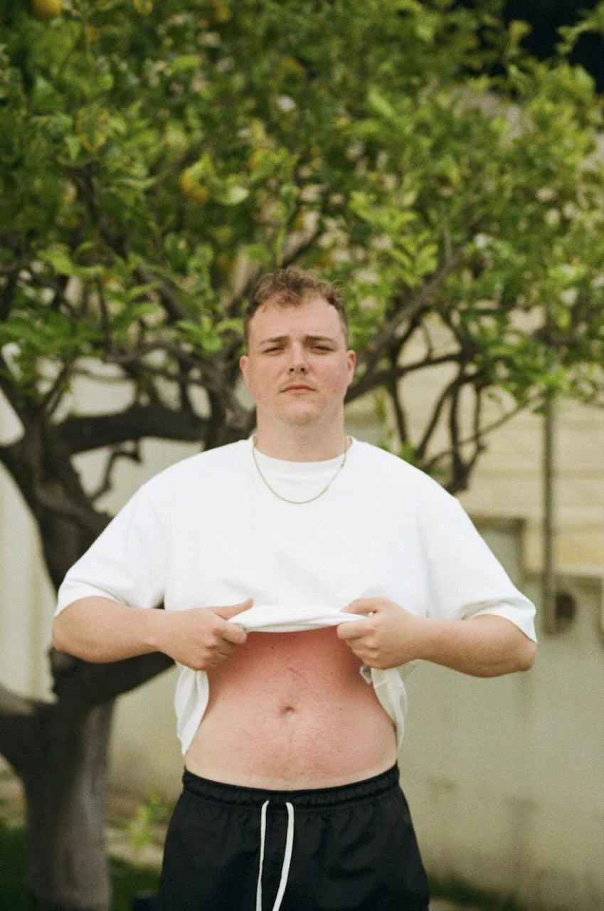 Junge mit weißen T-Shirt, das er hochzieht, um seinen Bauch zu zeigen, im Hintergrund ein Baum mit grünen Blättern.